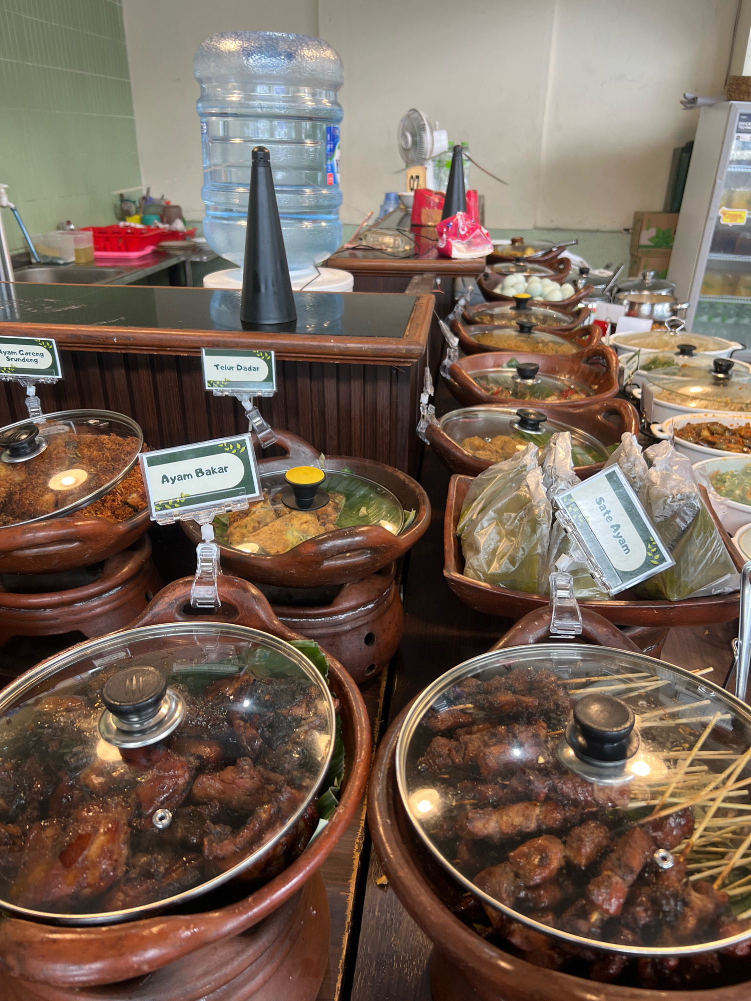 The image depicts a buffet-style food display, likely in a restaurant in Yogyakarta, Indonesia, given the Indonesian food labels. In the foreground, several clay pots with glass lids contain various dishes. One pot on the left holds what appears to be fried chicken with crispy toppings. Adjacent to it, a pot labeled "Ayam Bakar" (grilled chicken) displays grilled chicken pieces. To the right, a pot labeled "Sate Ayam" (chicken satay) contains several skewers of satay, some wrapped in banana leaves. Further back, other clay pots and serving dishes hold more food items, including what looks like hard-boiled eggs and other saucy dishes. A large water dispenser is visible in the background, along with a counter and what seems to be a refrigerator. The overall environment suggests a casual dining setting where customers can select their food.