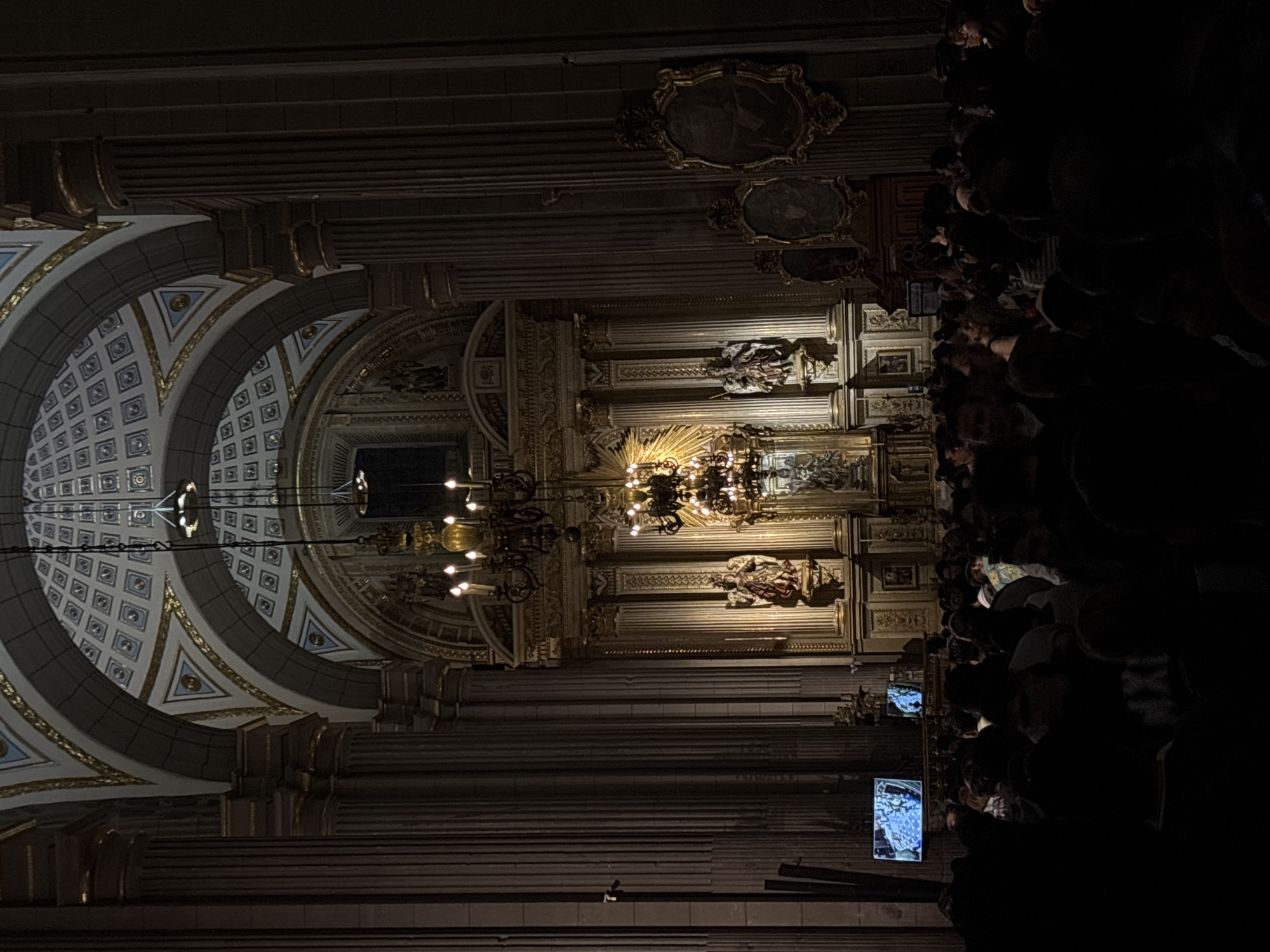The image captures the interior of the Puebla Cathedral in Puebla, Mexico. The shot is taken from the ground, looking up at the ornate vaulted ceiling, featuring intricate patterns and golden accents. A large chandelier hangs centrally, providing illumination.
Along the right side of the image, a crowd of people is visible, some holding up phones to take pictures or videos. The lighting is dim overall, emphasizing the details of the cathedral's architecture. Columns are prominent on the sides.
