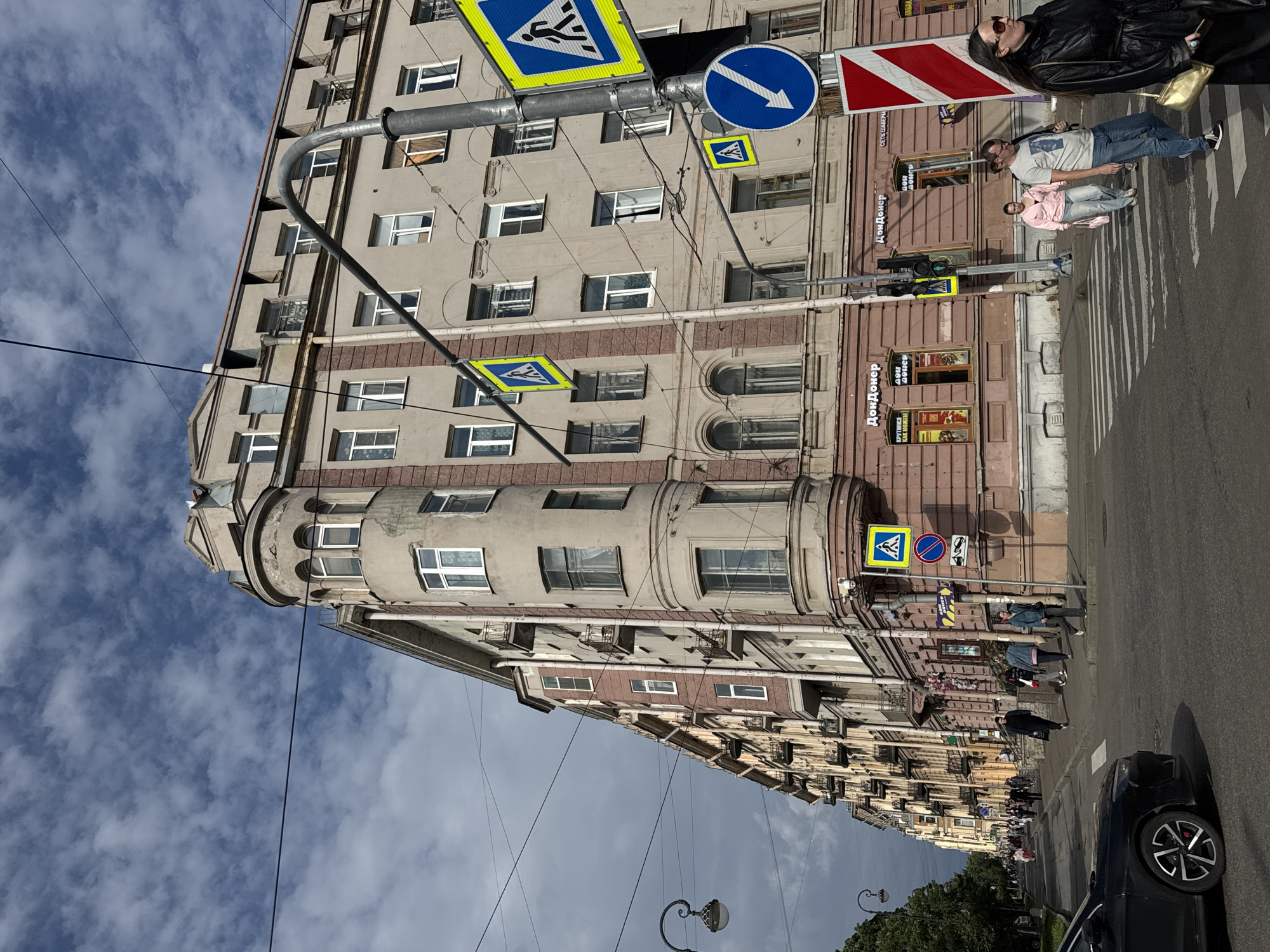 The image shows a street scene in Saint Petersburg, Russia, with a focus on an intersection and the surrounding buildings. The architecture is characteristic of the city, with multi-story buildings featuring ornate details and balconies. The sky is partly cloudy, indicating a typical day in Saint Petersburg.
At the intersection, several pedestrians are crossing the street. One woman in the foreground is wearing a black leather coat and sunglasses, carrying a gold purse. To her left, a man in a grey t-shirt and blue jeans is also crossing, looking towards the left. Behind him, a woman in a pink jacket and jeans walks with a pink bag.
Traffic signals are visible, including a pedestrian crossing light and a directional arrow. Several pedestrian crossing signs are posted on lampposts, indicating designated areas for crossing. The street is paved, and a black car is visible on the left side of the image, parked or driving slowly.
The buildings flanking the street are primarily commercial, with signs indicating businesses such as "ДонДонeр" (DonDöner), a fast-food establishment. The buildings are painted in various shades of cream and pink, with arched windows and decorative elements. Electrical wires crisscross the sky, a common feature in urban environments. The overall impression is a bustling city street on a moderately cloudy day.