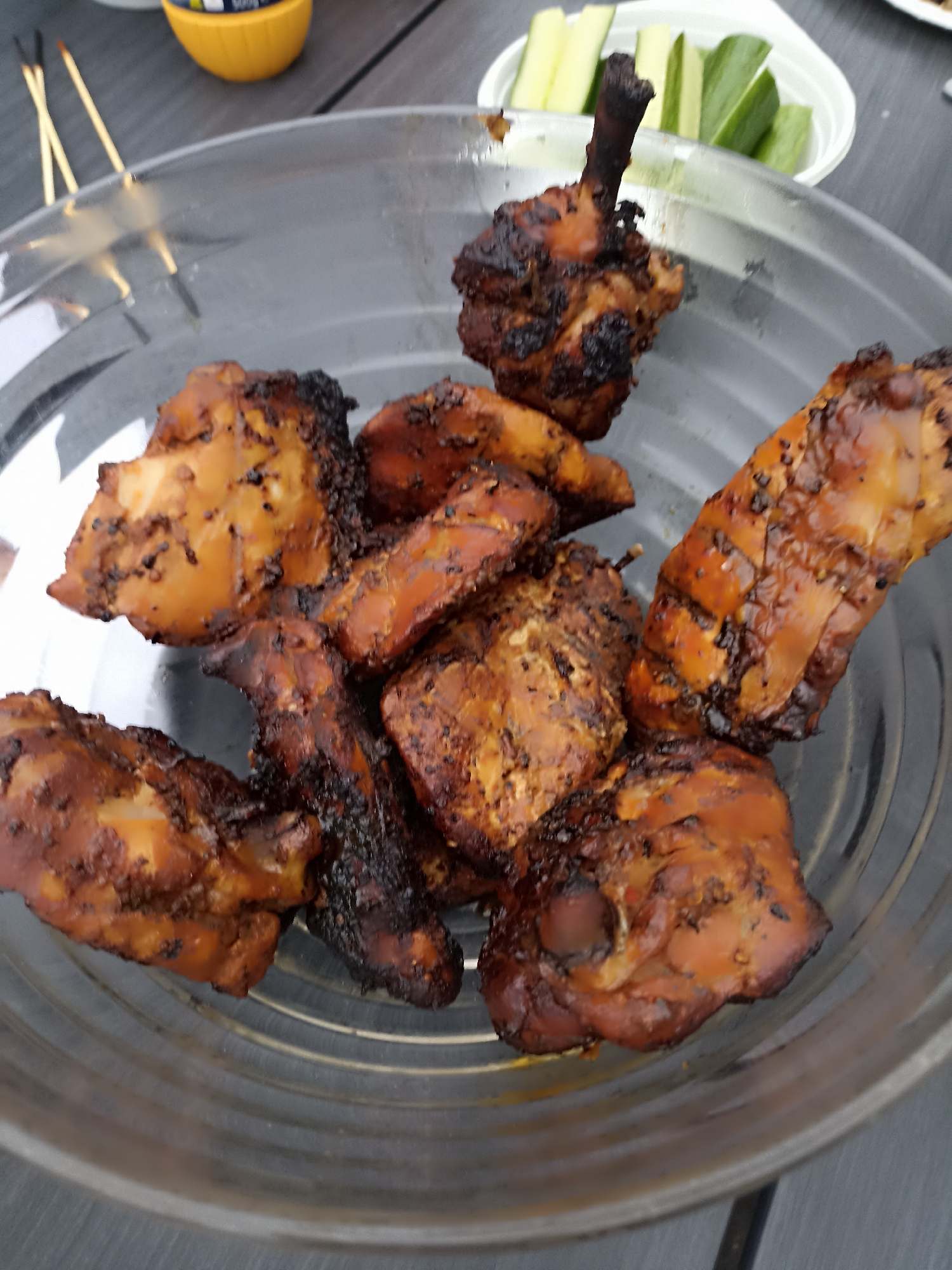 A clear glass bowl holds several pieces of grilled chicken. The chicken appears to be dark brown and glazed, with some charring. In the background, a white bowl contains sliced cucumbers. To the left, two wooden skewers are visible. The scene is set outdoors on a gray, textured surface, possibly a table. The lighting suggests it is daytime. The image shows food that is likely prepared for consumption. There is no discernible information about the location in Annagry, Ireland.