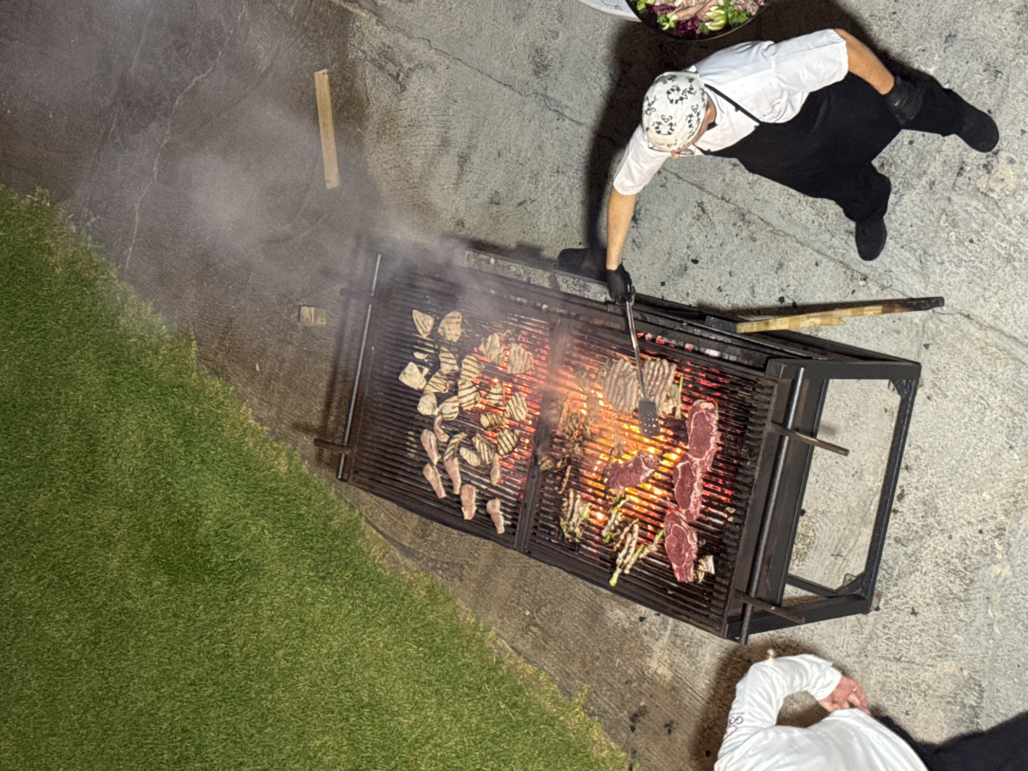 An aerial shot shows a chef grilling meat over a large, industrial barbecue in Isola delle Femmine, Italy. The barbecue has multiple levels of grates laden with various cuts of meat, some appearing to be steaks and others smaller, thinner pieces. Flames are visible beneath the meat, indicating intense heat and cooking. Smoke rises from the grill, mingling with the night air. The chef, wearing a white shirt, black apron, and a patterned bandana, uses tongs to tend to the food. To the left of the barbecue, a person in a white shirt stands with their hand near their waist. The scene is set on a concrete patio adjacent to a neatly mowed grassy area. The overall impression is one of an outdoor culinary event taking place after dusk.