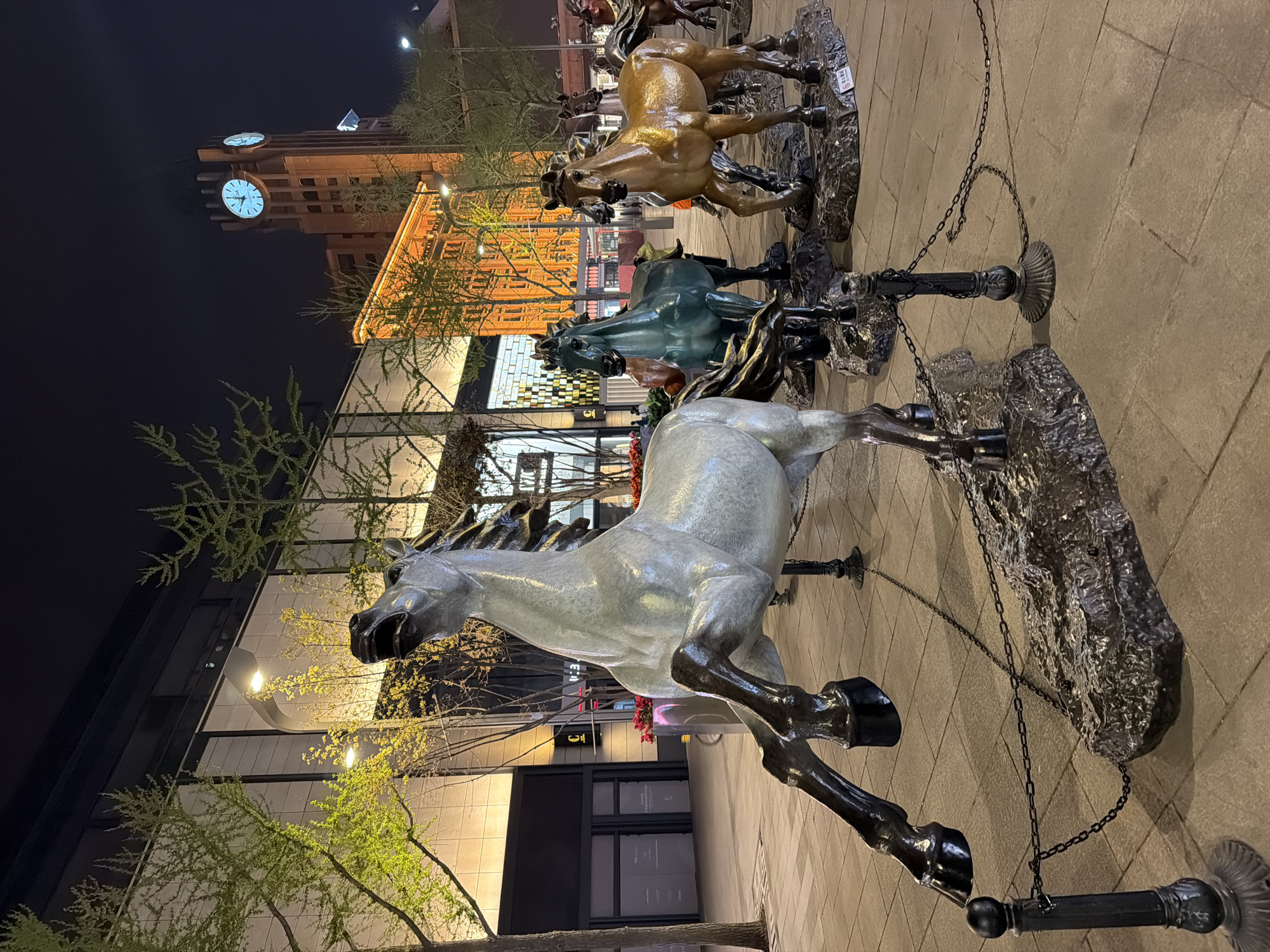The image depicts a statue of galloping horses at night. The statue appears to be made of bronze or a similar material, with a white horse in the foreground and several other horses of different colors behind it. The horses are mounted on rocky bases, with chains connecting them.
The background includes a building with a clock tower, illuminated at night. The style of architecture suggests an urban setting.
Based on the location in the city of Dongcheng District, China, it may be a depiction of the "Eight Horses" statue which is located in Wangfujing pedestrian street.