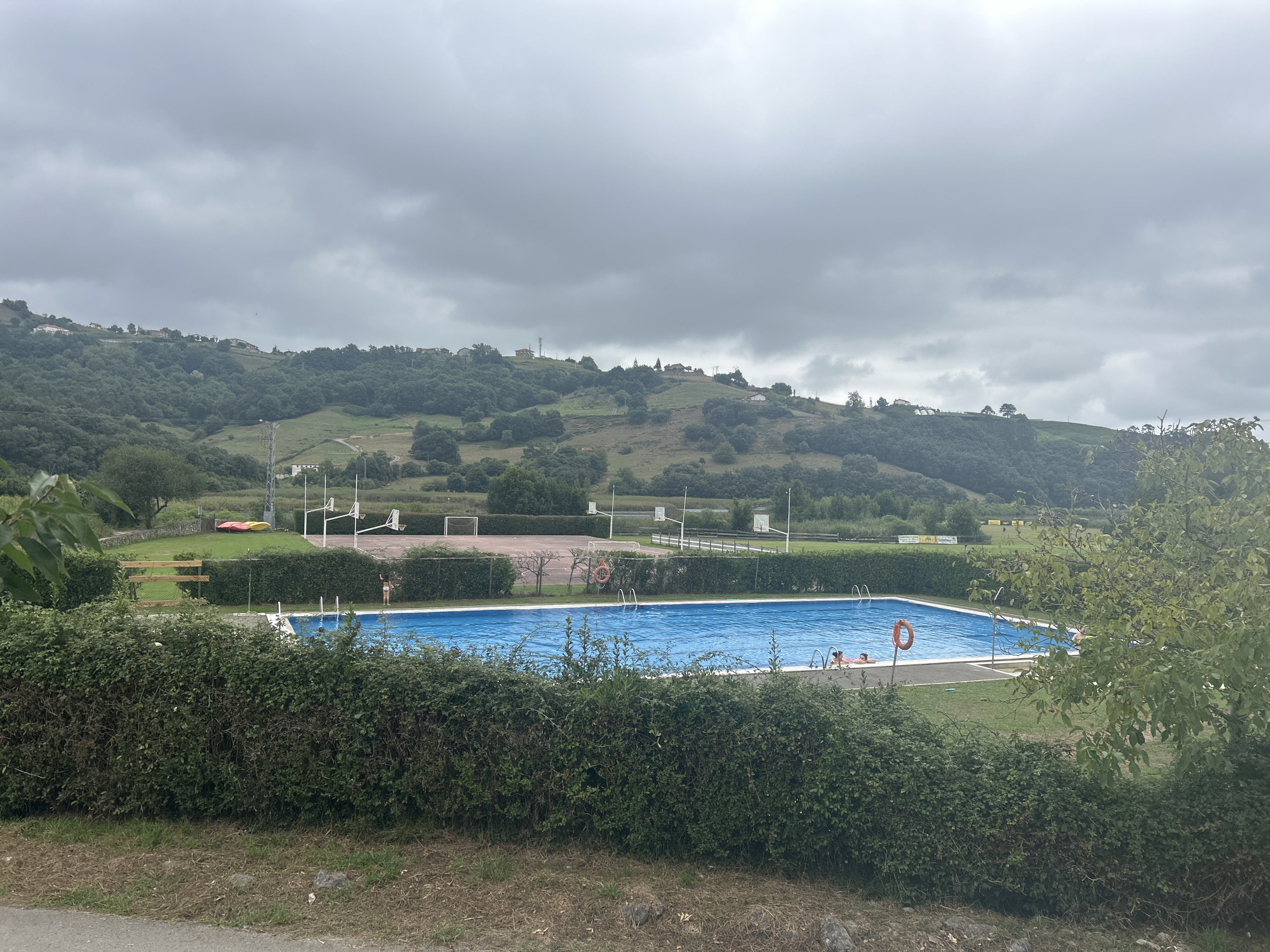 The image depicts an outdoor swimming pool facility in Voto, Spain. The pool itself is filled with bright blue water and has several people visible in and around it. On the right side of the pool, two individuals are partially submerged, seemingly relaxing. There are also ladders providing access to the water. A bright orange life preserver ring is placed on the edge of the pool. Surrounding the pool is a neatly trimmed green hedge. Beyond the hedge, to the left of the pool, is a grassy area with a wooden bench and a fence. Further in the background, there are sports courts with basketball hoops and a soccer goal. The entire facility is set against a backdrop of lush green hills covered with trees and dotted with small houses and buildings, characteristic of the Voto landscape. The sky is overcast with heavy grey clouds, suggesting a cloudy day. A road or path is visible in the foreground, with dry grass and some rocks on its edge.
