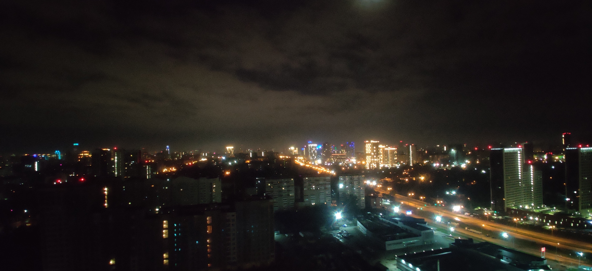 This image is a nighttime aerial view of a cityscape in Yekaterinburg, Russia. The scene is dominated by the dark sky, with clouds partially obscuring a visible moon. Below, a sprawling urban area is illuminated by a multitude of lights from buildings and streetlights.
In the foreground, several multi-story residential buildings are visible, some with illuminated windows showing warm yellow and cool blue light. These buildings appear to be apartment blocks with consistent architectural styles.
Further into the mid-ground and background, the cityscape becomes denser with taller, modern buildings. Some of these structures are brightly lit with decorative colored lights, featuring blue and white accents, suggesting they might be commercial or office buildings. A prominent cluster of these illuminated towers stands out, with one building exhibiting a repeating pattern of bright yellow lights. Another tall building in the distance has red lights at its apex.
A major road or highway cuts through the city, indicated by the bright streaks of headlights and taillights from moving vehicles. Streetlights line the road, casting a warm orange glow. The presence of cars and the illumination of the road suggest active traffic flow.
The overall impression is a dynamic and illuminated urban environment at night, characteristic of a large city. The specific location within Yekaterinburg is suggested to be an area with a mix of residential and modern commercial architecture, likely a developing or established urban district. No individuals or specific actions are discernible due to the distance and nighttime conditions.