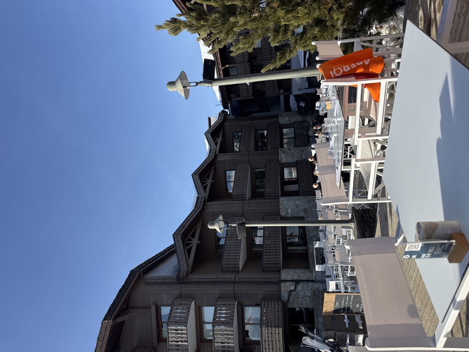The image depicts an outdoor restaurant terrace in Courchevel, France, under a clear blue sky. In the foreground, a white square table is visible, featuring a small wooden stand displaying a "PAY THE BILL" sign with a QR code. Numerous other white tables and chairs are arranged across the terrace, which appears to be a wooden deck. At a mid-ground table, an orange "APEROL" branded towel hangs over a white chair, and several clear glasses are set on the table surface. Multiple individuals are seated at various tables in the mid-ground, engaged in outdoor dining or socializing. Two tall streetlights with grey, bell-shaped shades are positioned within the terrace area. In the background, large, multi-story chalets constructed from dark wood paneling and stone bases are visible, featuring multiple balconies with wooden railings and windows reflecting the sky. The roofs of these buildings are sloped and show patches of snow. A tall green coniferous tree stands on the right side, partially obscuring a building. On the far left, a pair of skis leans vertically against a dark structure, and a dark chalkboard or menu stand is present near the edge of the terrace. Patches of snow are visible on the ground beyond the terrace perimeter.