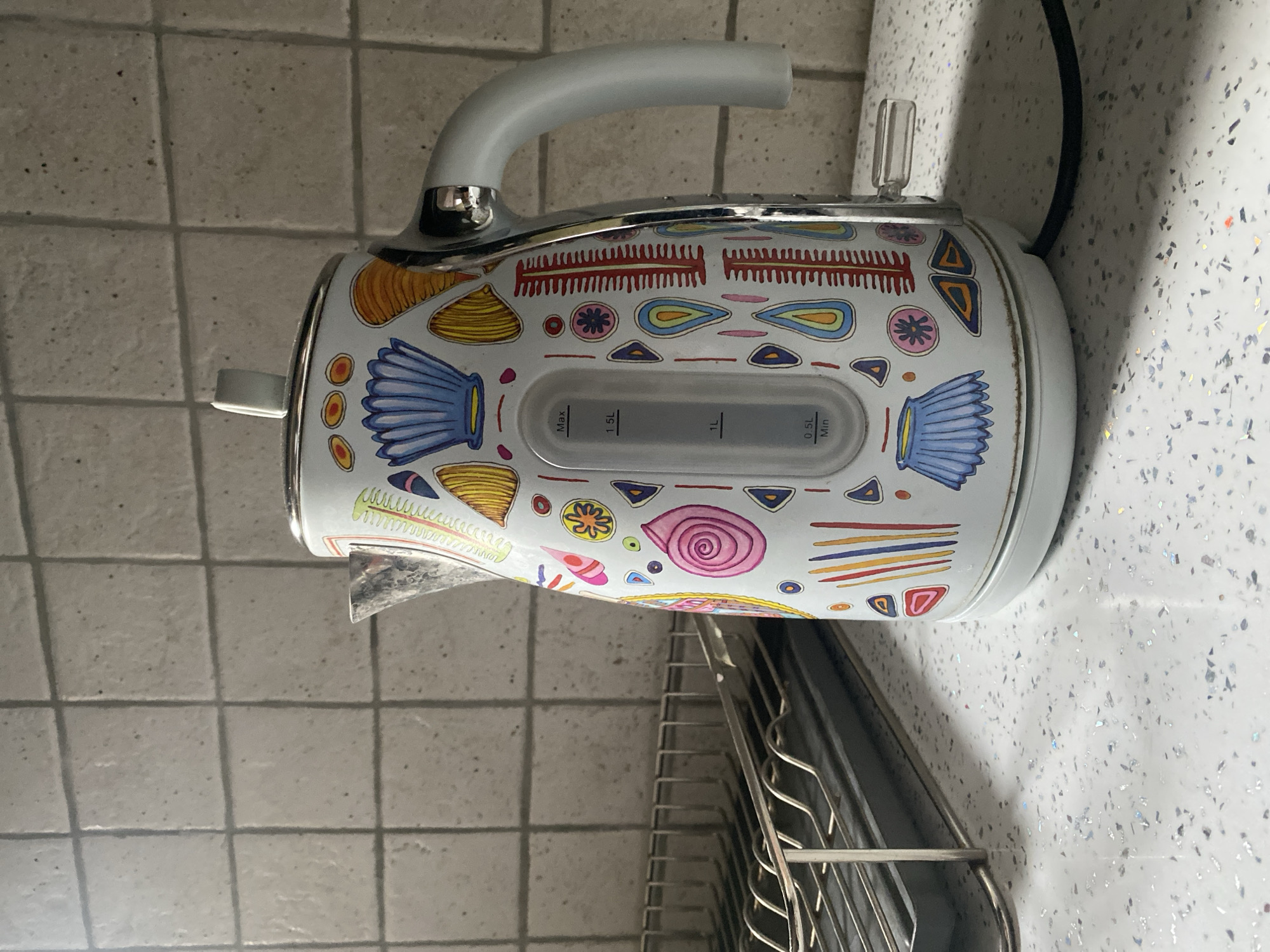 The image shows a white electric kettle with a colorful, artistic pattern of abstract shapes and designs. The kettle has a silver spout and handle, and a clear water level indicator with markings for 0.5L, 1L, and 1.5L. It is placed on a speckled white countertop, next to a metal dish rack. In the background, a light-colored tiled wall is visible. The lighting suggests an indoor setting, possibly a kitchen. The overall scene is a still life of kitchenware.