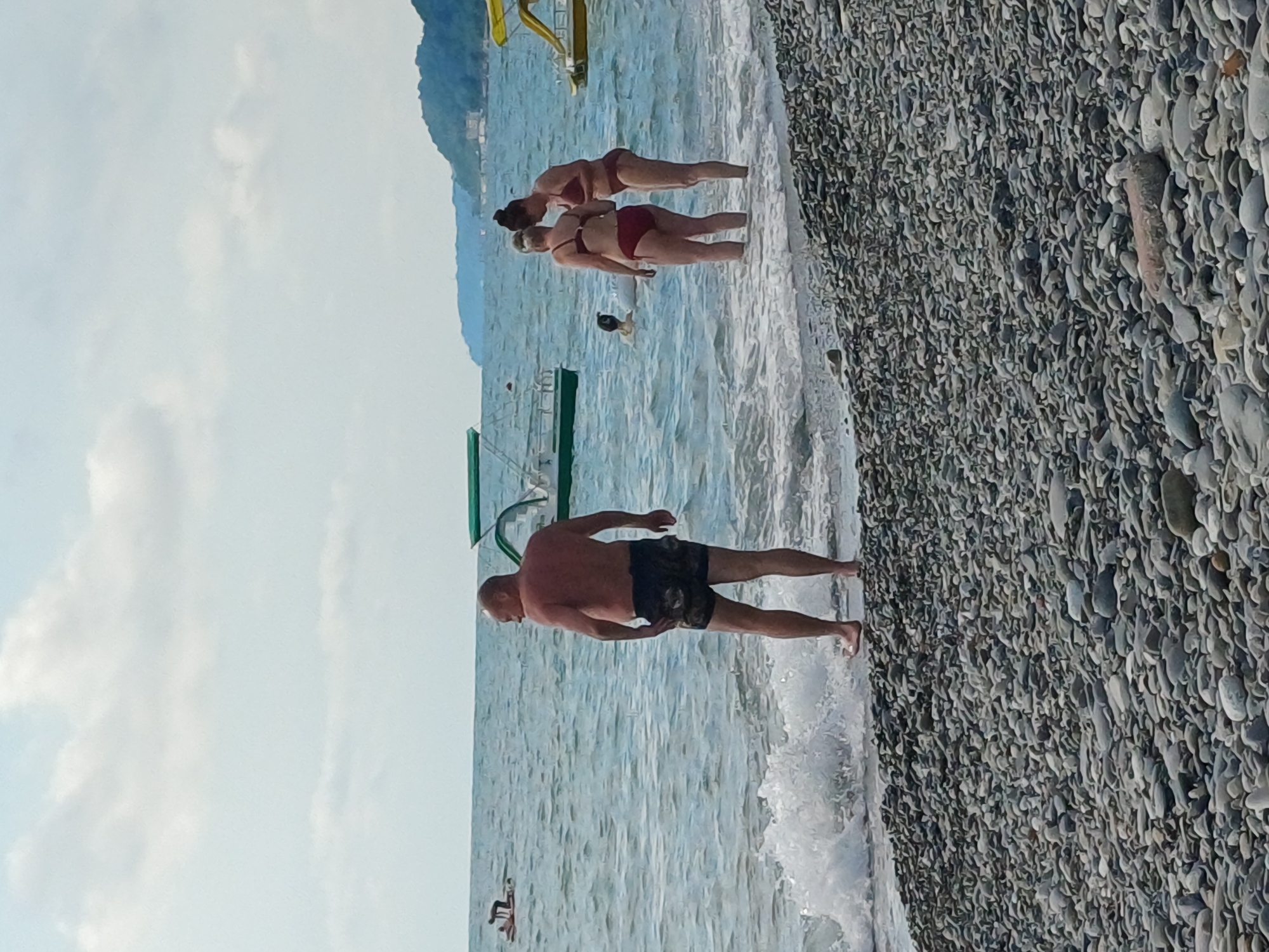 In Sochi, Russia, on a cloudy day, a pebbled beach meets the calm sea. A man in dark patterned shorts stands facing away from the viewer, his back to the ocean. In the water, two women in red bikinis are wading, one with her back to the viewer and the other facing her. Further out, a person is partially visible in the water. Two boats are moored in the sea; one is a green and white boat with a canopy, and another, yellow and white boat with a canopy is further to the right. In the distance, a tree-covered mountain slopes down to the coast.