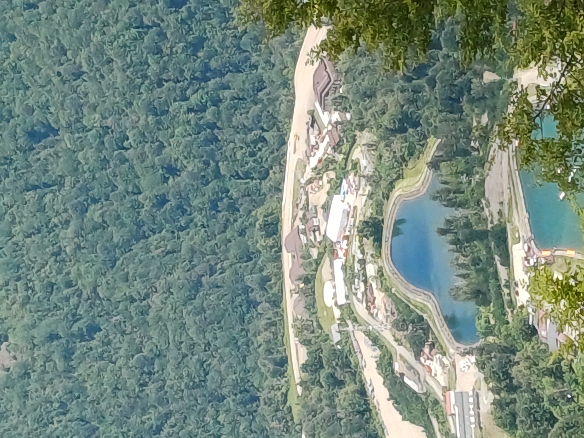 This aerial photograph captures a view of the Estosadok, Russia area, showcasing a settlement nestled amongst lush, green forested mountains. A prominent feature is a body of water with a distinctive heart shape, possibly a reservoir or a decorative pond, with a manicured embankment surrounding it. Nearby, a wider, muddy river snakes through the landscape. The settlement itself consists of numerous buildings, including what appear to be industrial or commercial structures with light-colored roofs, as well as some residential-looking buildings with darker, sloped roofs. Roads and pathways connect the various parts of the settlement, and some areas show signs of construction or land clearing. The dense forest covers the surrounding hillsides, indicating a natural, undeveloped environment adjacent to the developed area. The lighting suggests it is daytime with ample sunlight. There are no visible people in the photograph.