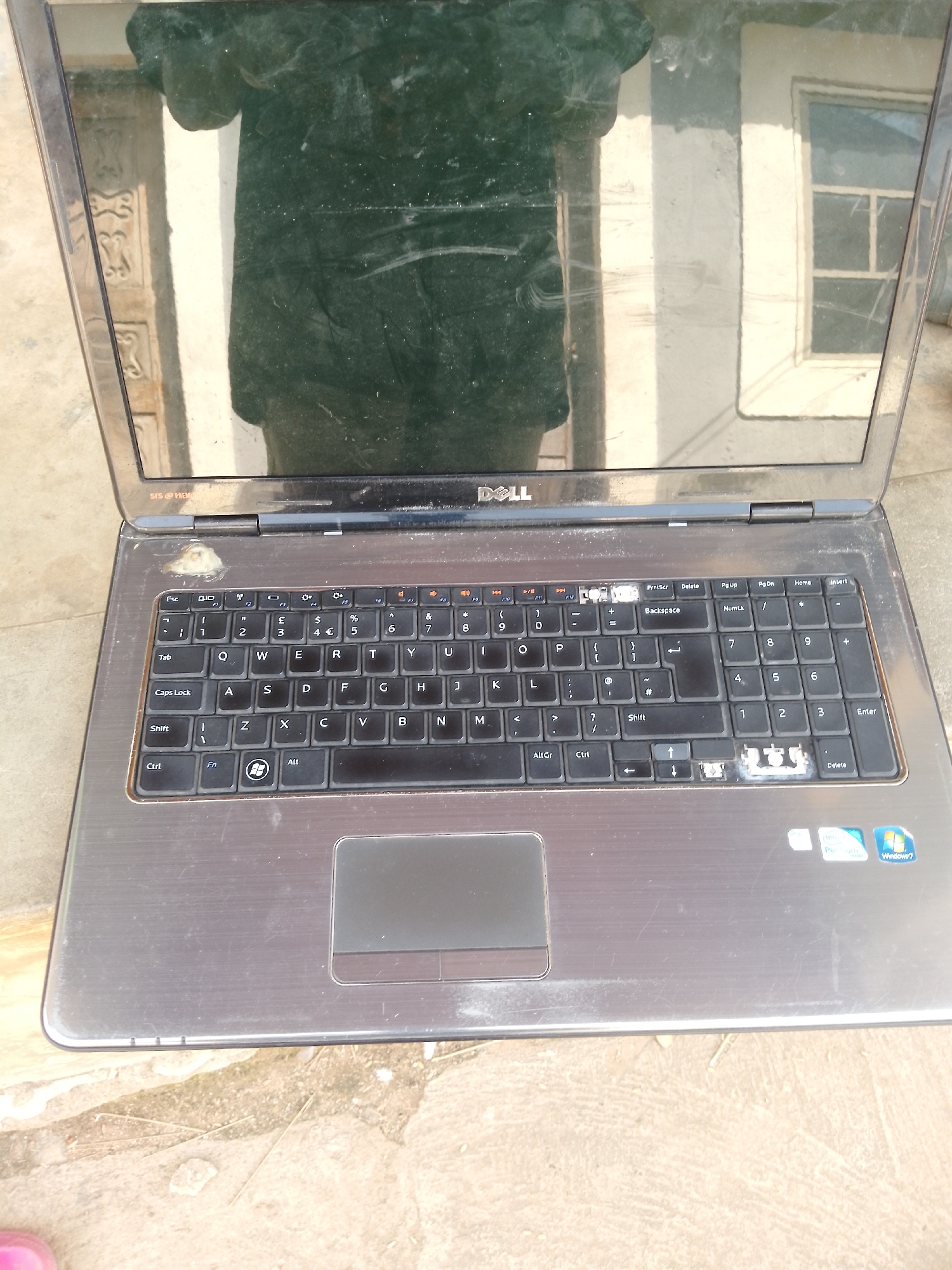 This is a close-up, top-down view of an open, used Dell laptop computer placed outdoors on a rough, dusty ground. The laptop's screen is reflecting a dark, blurry image of what appears to be a person and a building with windows. The keyboard is visible, showing a standard QWERTY layout with some keys missing or damaged, particularly on the right side of the numeric keypad. The trackpad is also visible. There are various stickers on the laptop's casing, including one that says "Windows 7". The overall condition of the laptop suggests it has been used extensively and is in a state of disrepair. The environment appears to be a rural or semi-urban setting in Serti, Nigeria, characterized by the rough ground and outdoor placement. There are no people directly visible in the image, but the reflection in the screen suggests the presence of at least one individual. The lighting is natural, indicating it is daytime.