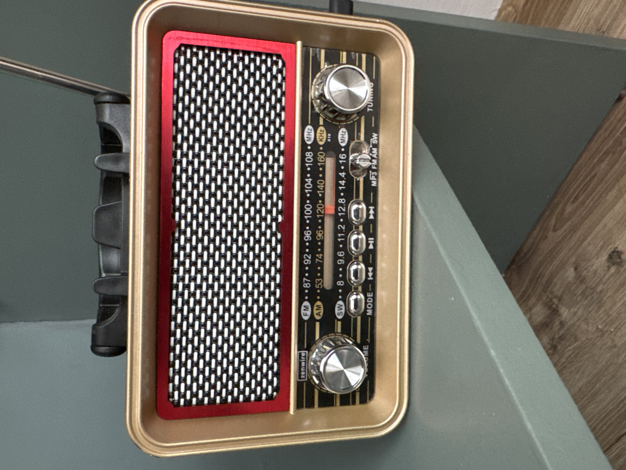 The image depicts a Zenwire radio with a gold-colored frame, featuring a red grille and two silver tuning knobs. The radio is positioned on a gray surface with a visible wood-like floor in the background. The controls indicate it has FM, AM, and SW bands, along with MP3 playback capability. A black antenna is partially visible on the left side of the frame. The location is unspecified.
