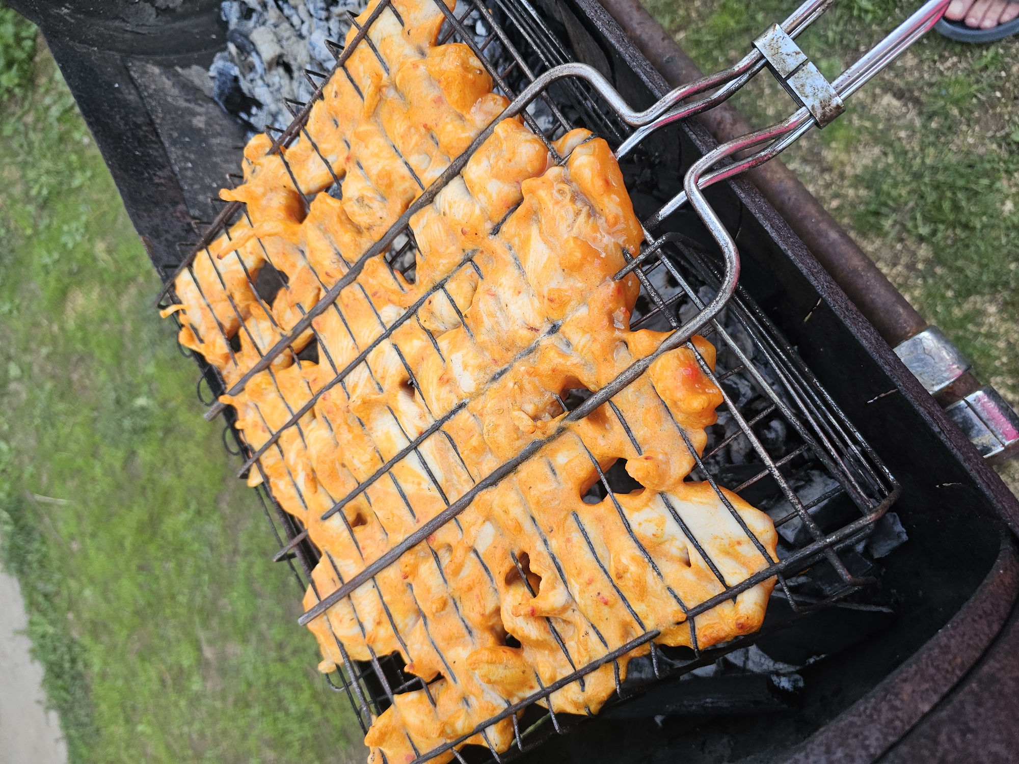 A high-angle outdoor shot shows chicken coated in a bright orange sauce being grilled in a metal basket over charcoal. The grill is part of a larger barbecue, likely located in a backyard or park in Альняш, Russia. Green grass is visible in the background. A person's foot wearing a black sandal is visible in the upper right corner. The focus is on the chicken cooking in the basket over the grill.