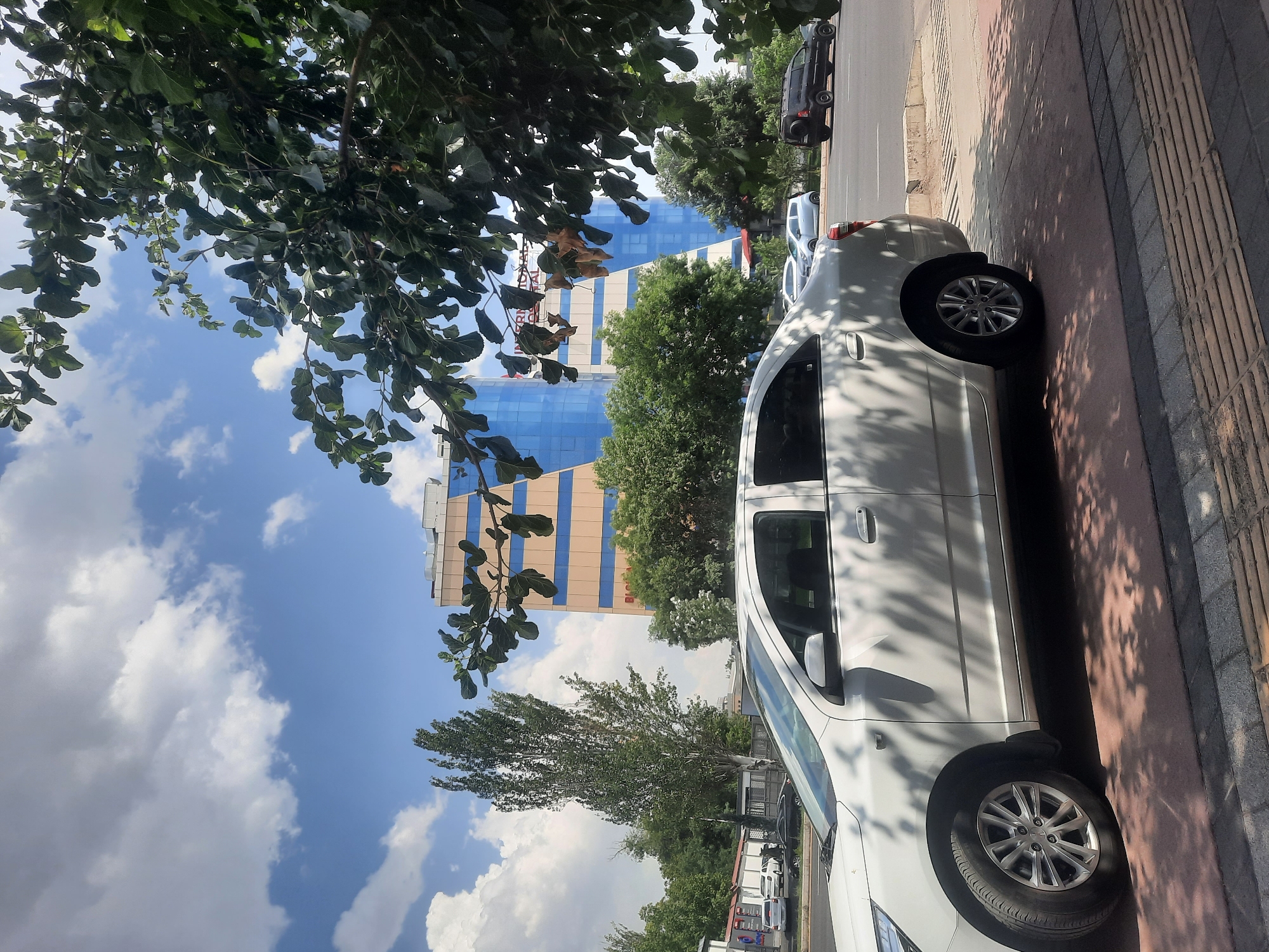 A white sedan is parked on the side of a road in Tashkent, Uzbekistan, on a sunny day with scattered clouds. The car is positioned in the foreground, taking up a significant portion of the frame. Beyond the car, a multi-story building with a blue glass facade and beige and orange accents stands prominently. Trees and foliage are visible throughout the scene, with some branches and leaves casting shadows on the car and the ground. In the background, other vehicles, including a black SUV and a light-colored hatchback, are on the road. The area appears to be an urban setting with a sidewalk and a paved road.