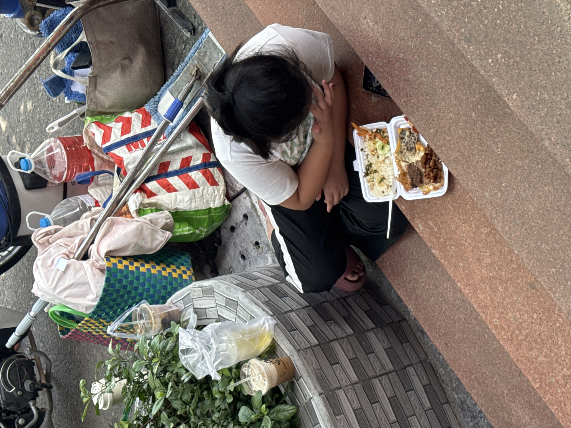 In the city of Bac Nha Trang Ward, Vietnam, a young individual with dark hair, wearing a white t-shirt and black pants with a white stripe, sits on reddish-brown granite steps. Their body is hunched over, with arms crossed and head lowered, appearing to be resting or possibly eating. In front of them, on the steps, is a white Styrofoam container filled with food, including rice, what appears to be fried meat, and some vegetables. To the left of the individual and further up the steps, a collection of items is gathered. These include several plastic cups with straws, one containing a brown beverage, and another with a yellow liquid. A large green and yellow woven basket is visible, filled with light-colored fabric, likely clothing. A red and white striped bag is also present, along with cleaning supplies such as a mop with blue and white cloths. A partially visible motorcycle is to the far left, and lush green foliage borders the area to the left of the steps. The ground appears to be asphalt, and the overall scene suggests a temporary resting spot amidst daily activities.