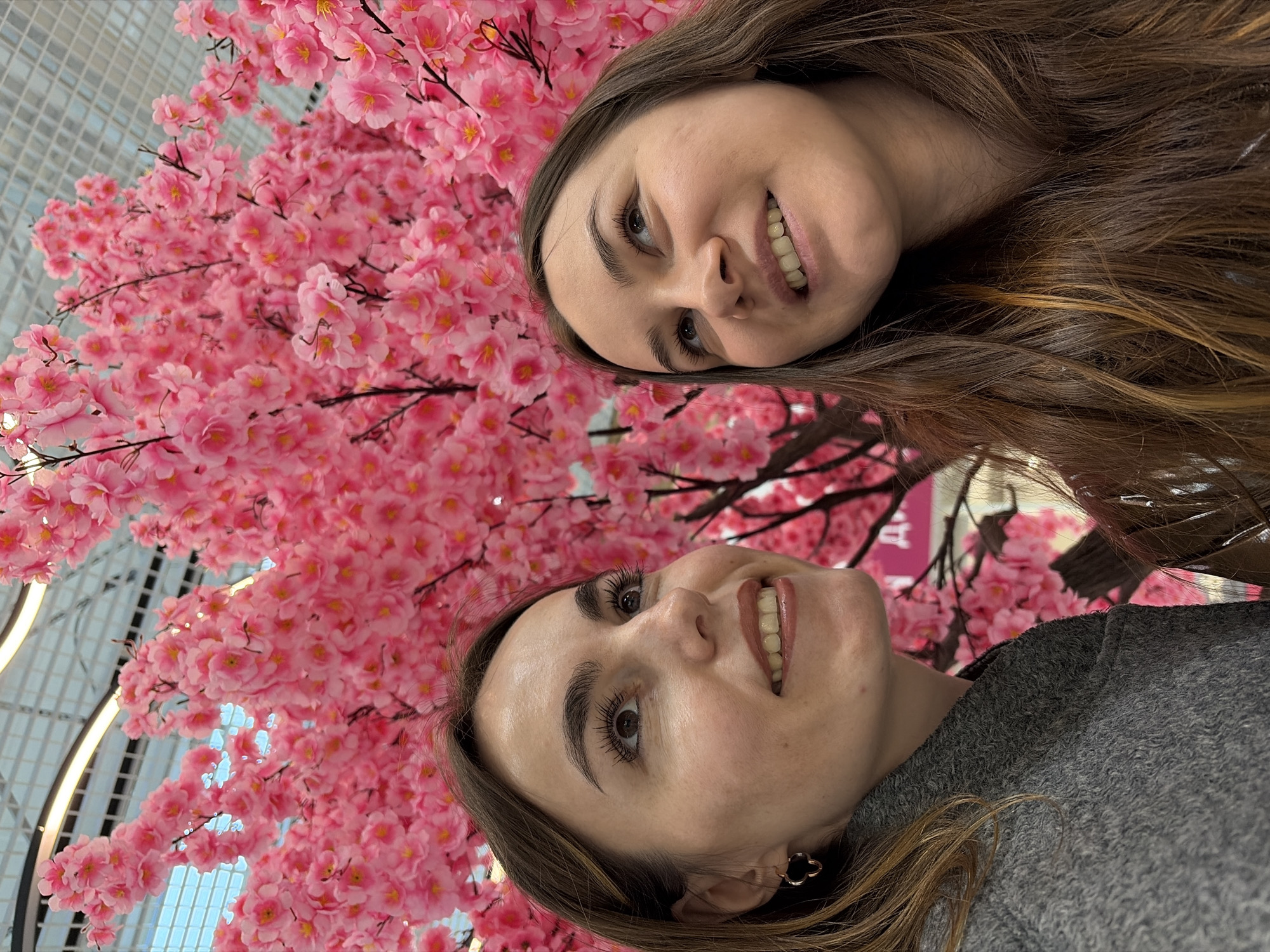 Two women are posing for a picture with a faux cherry blossom tree in what appears to be an indoor location. One woman is wearing a gray top, while the other woman's attire is not fully visible. The background shows a grid-like ceiling with lighting fixtures, suggesting an indoor setting that might be a shopping mall or public space. The image's perspective is slightly tilted. The pink cherry blossoms are a prominent feature, creating a cheerful and vibrant backdrop. Based on the hint, the location might be in the city of Izhevsk, Russia.