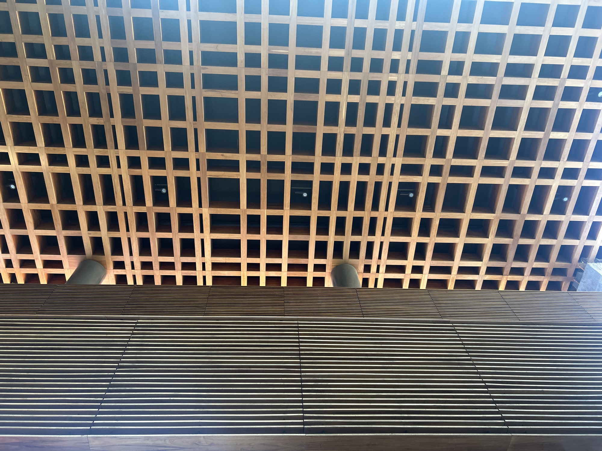The image shows a low-angle view of a building interior featuring distinctive architectural details. The bottom portion of the frame is filled with a horizontally aligned facade composed of numerous thin, parallel slats, illuminated to create a high-contrast light and shadow pattern. Above this section, there's a transition to a three-dimensional wooden grid structure forming a decorative ceiling. The grid is composed of evenly spaced wooden beams, creating a pattern of squares. Within some of the grid squares are recessed lights. Two gray cylindrical objects, potentially support columns or ventilation shafts, intersect with the transition point between the facade and the grid. The overall impression is a modern architectural design emphasizing geometric patterns and contrasting materials. The location is provided as Unknown, Unknown.