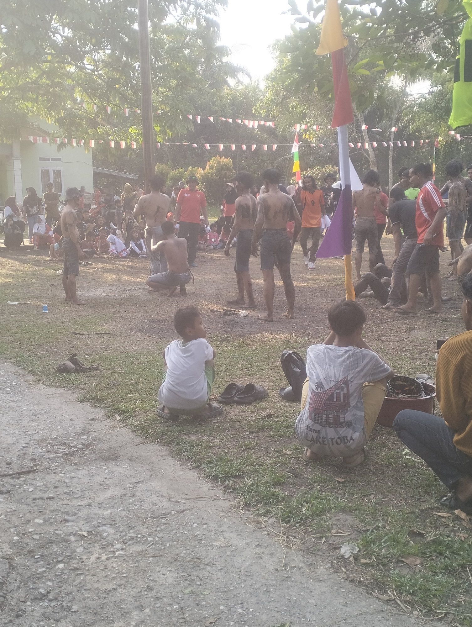 The image depicts an outdoor scene in the city of Dumai, Indonesia, possibly during a cultural event or celebration. The ground is a mixture of dirt and sparse grass. Several people, predominantly young men, are visible. Many of them have their torsos bare and appear to be covered in mud, suggesting participation in a ritual or activity involving dirt.
In the foreground, two young boys are seated on the ground. One boy is wearing a white t-shirt and green shorts, facing away from the camera. The other boy, also facing away, is wearing a grey t-shirt with a graphic print that includes the text "Horas LAKE TOBA", and yellow pants.
In the background, a crowd of people, including women and children, are gathered. Some individuals are standing, while others are seated or crouching. Decorative flags, including red and white Indonesian flags and brightly colored flags, are strung across the area, contributing to a festive atmosphere. A building with white walls and a green roof is visible in the far background, along with trees and vegetation. The lighting suggests it is daytime, with sunlight filtering through the trees. The overall impression is one of a community gathering, likely associated with a local tradition or event.