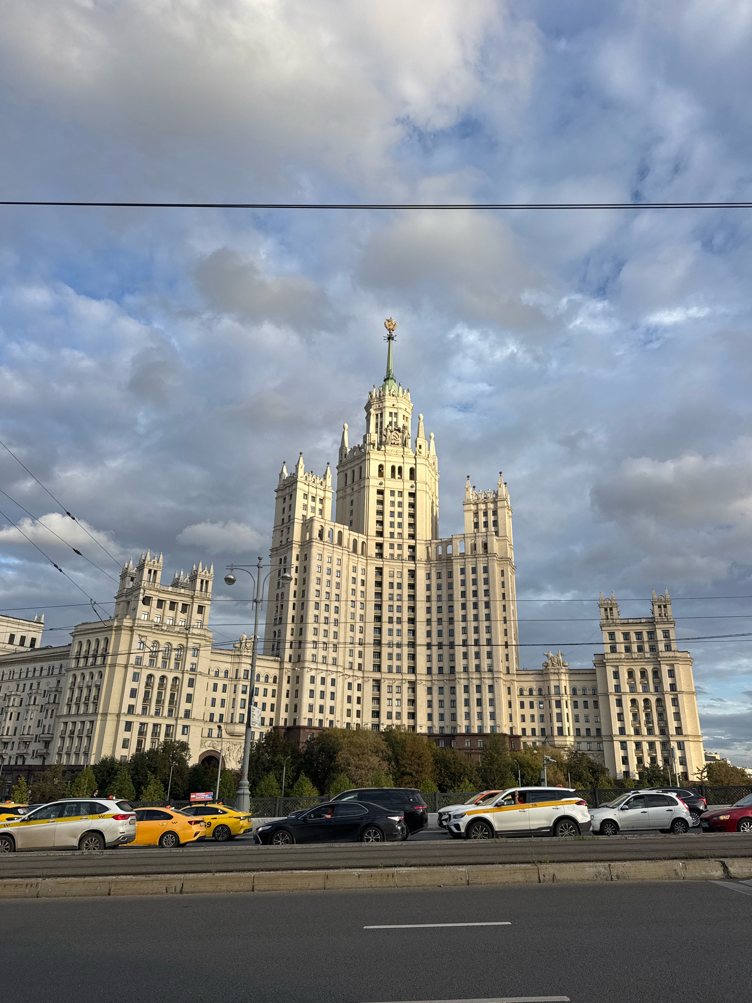 This is a full-shot, low-angle, exterior, eye-level, natural light photograph of the Kotelnicheskaya Embankment Building, a Stalinist skyscraper in Moscow, Russia, located at the confluence of the Moskva River and Yauza River. The building, constructed between 1947 and 1953, is a prominent example of Stalinist architecture, characterized by its imposing scale, ornate detailing, and a central spire topped with a star. The building's facade is light-colored and features numerous windows, balconies, and decorative elements, including smaller towers and turrets. The sky is partly cloudy with dramatic, dark clouds contributing to a moody atmosphere. In the foreground, a multi-lane road is visible with several cars and taxis in traffic. The road surface is grey asphalt, and a concrete barrier separates it from the area where the building stands. The architecture of the building dominates the background, with trees and greenery visible between the road and the structure. Overhead, power lines are present, crisscrossing the sky. The overall scene depicts a busy urban environment with a historic and iconic skyscraper as the focal point.