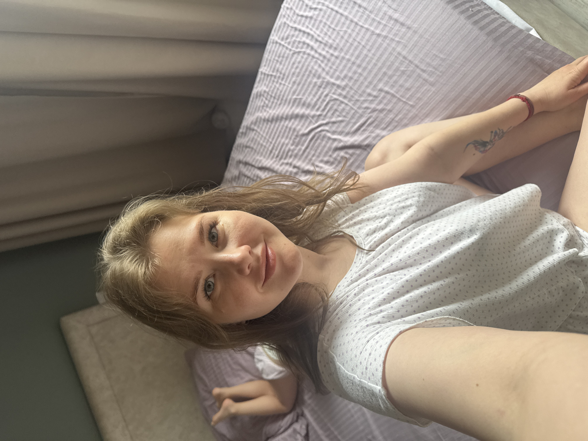 A fair-skinned woman with light brown hair and blue eyes is lying on a bed with purple striped bedding. She is wearing a white t-shirt with a small polka dot pattern. A red bracelet is visible on her right wrist, and a tattoo is on her right forearm. A small child's leg is visible in the lower left corner of the frame. The scene appears to be indoors, with a beige headboard and a gray wall visible. The location is unknown, but specified by the request as Gornyi (Dvurechenskoe S/P), Russia.