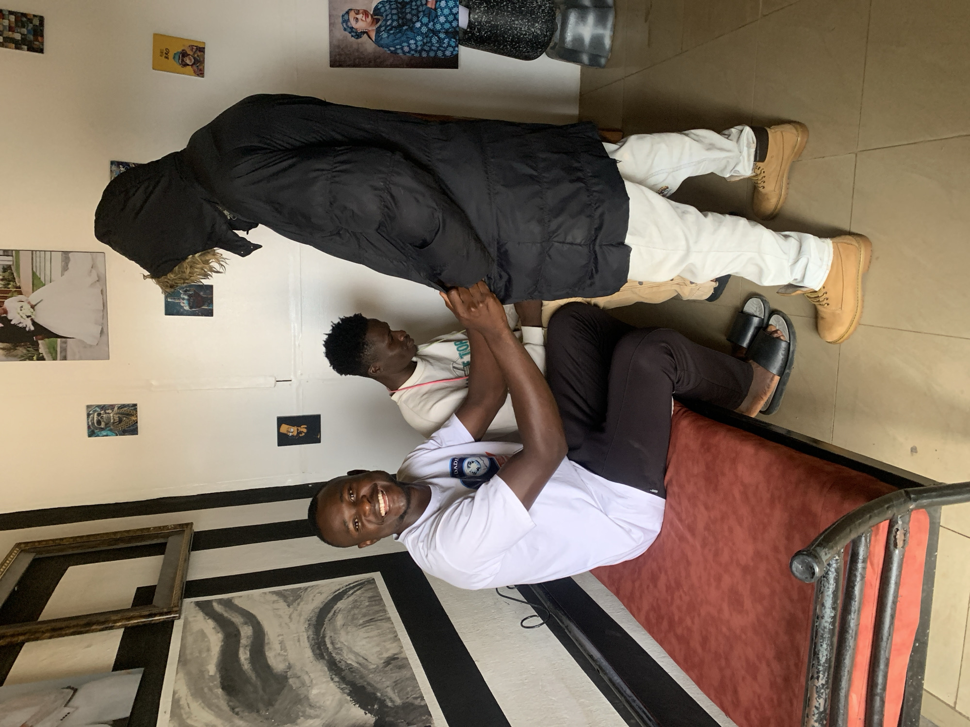 In a room in Jos, Nigeria, a Black man in a white t-shirt and black pants smiles broadly while sitting on a red bench. He is on the left side of the image. To his right, another Black man, wearing a white t-shirt and beige pants, is bending over and appears to be adjusting the zipper or pocket of a third Black man who is facing away from the camera. This third man is wearing a black hooded puffer jacket and light-colored pants. The room has striped black and white walls to the left, with artwork and framed pictures on the wall. The floor is tiled.
