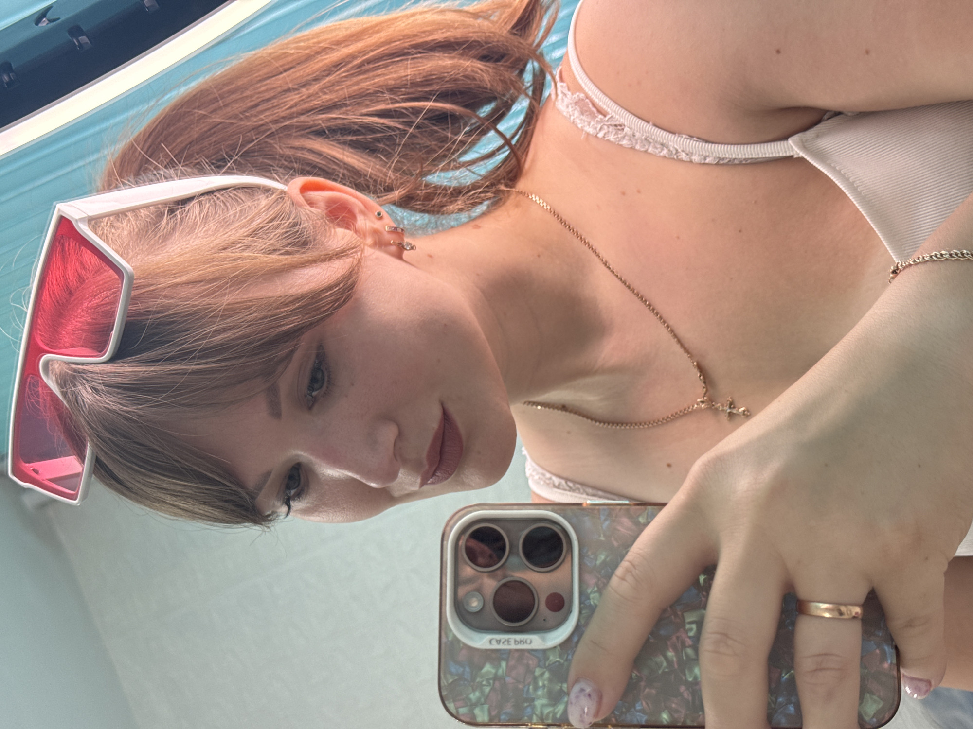 In the city of Chaikovsky, Russia, a young woman with auburn hair styled in a ponytail and bangs poses for a selfie. She wears white-rimmed sunglasses with red lenses pushed up onto her forehead. Her right hand holds a smartphone with a colorful, iridescent case. She is also wearing a delicate gold necklace with a cross pendant, a gold ring on her left ring finger, and a gold chain bracelet. Her top appears to be a ribbed, white camisole with lace trim. The background is a bright, teal-colored surface, possibly a wall or fabric, with a lighter blue section to the left. The lighting is bright, suggesting a well-lit indoor environment.