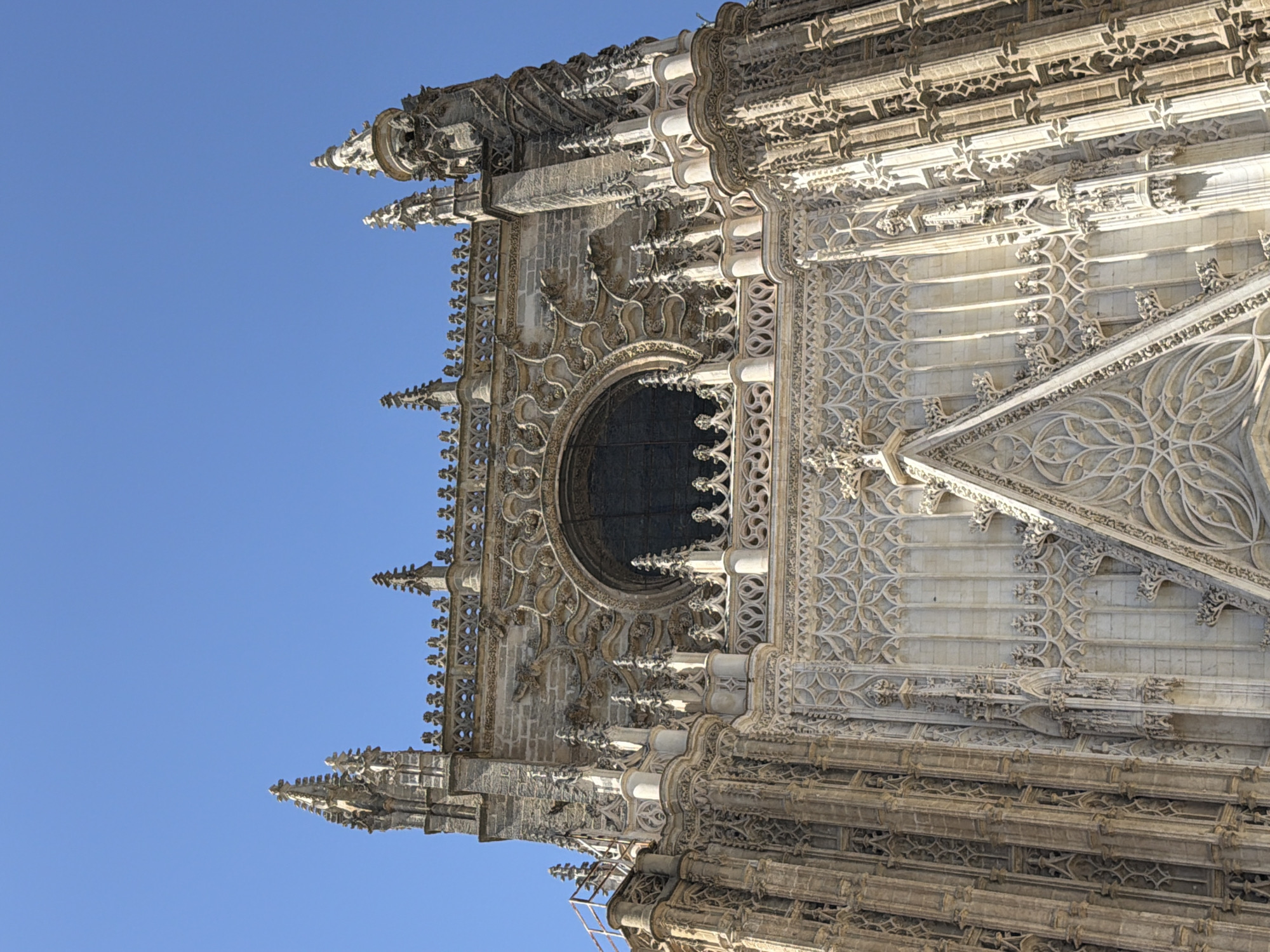 The image depicts a close-up view of the intricate Gothic architecture of a cathedral, likely Seville Cathedral in Spain, set against a clear blue sky. The structure is made of weathered stone, showcasing a wealth of detailed carvings, pointed spires, and decorative tracery. The sunlight suggests it is daytime, casting subtle shadows that highlight the depth and complexity of the stonework. The scene is static, with no people or immediate activity visible, focusing entirely on the architectural grandeur. Notable details include the repeating geometric patterns in the stonework, the delicate finials adorning the spires, and the arched window with its elaborate surround. The perspective is from below, looking upwards at the towering facade, emphasizing its scale and imposing presence. The overall impression is one of historical significance and artistic craftsmanship. There is no visible text in the image.