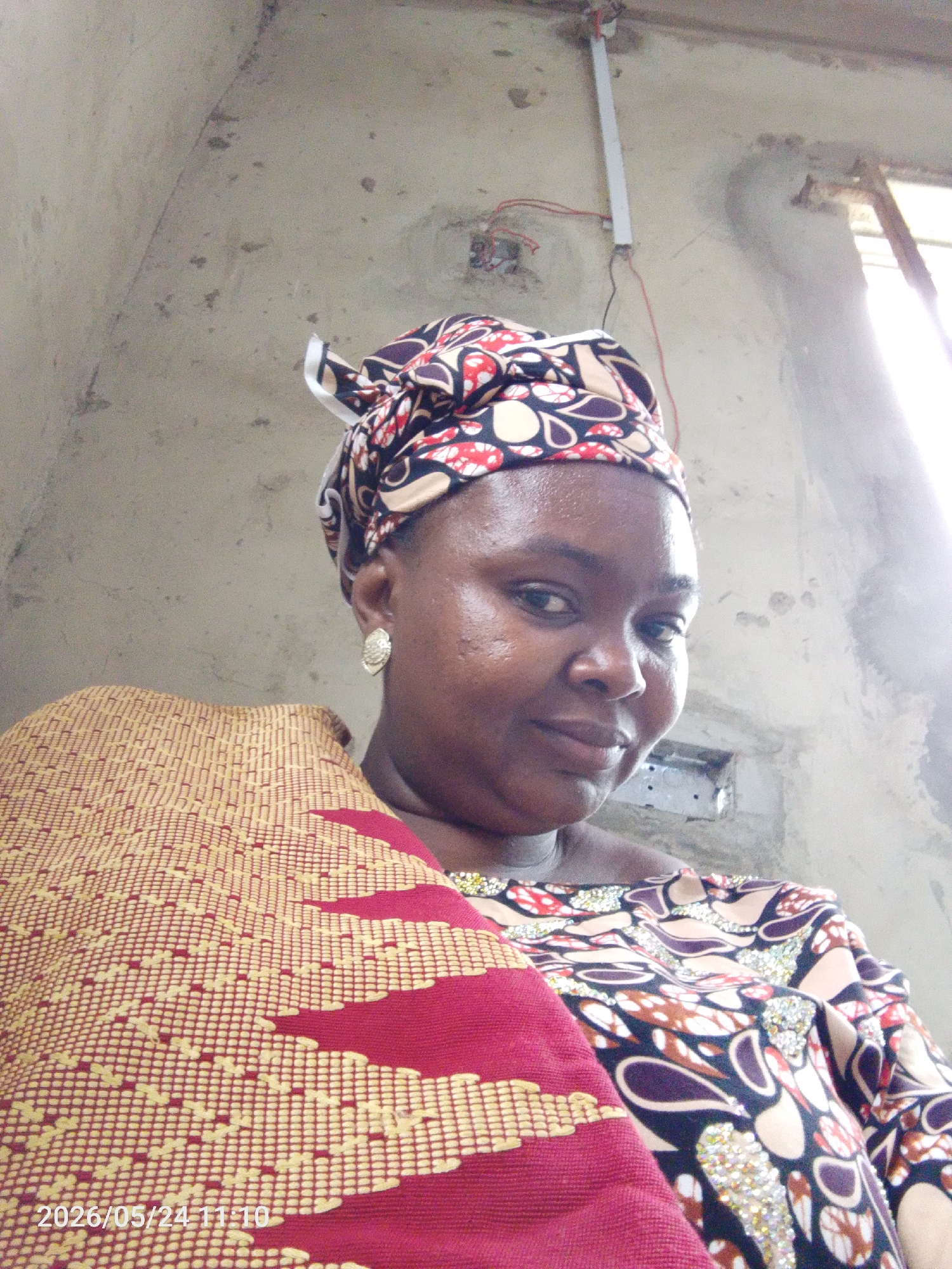 The image shows a person indoors, presumably in a building within Lokoja, Nigeria. The individual is wearing a patterned headwrap and a matching patterned top. A red and yellow woven blanket is draped over their shoulder. The walls are unfinished concrete, and electrical wiring is visible near the ceiling. A small window is also visible. The photo has a timestamp of "2026/05/24 11:10".