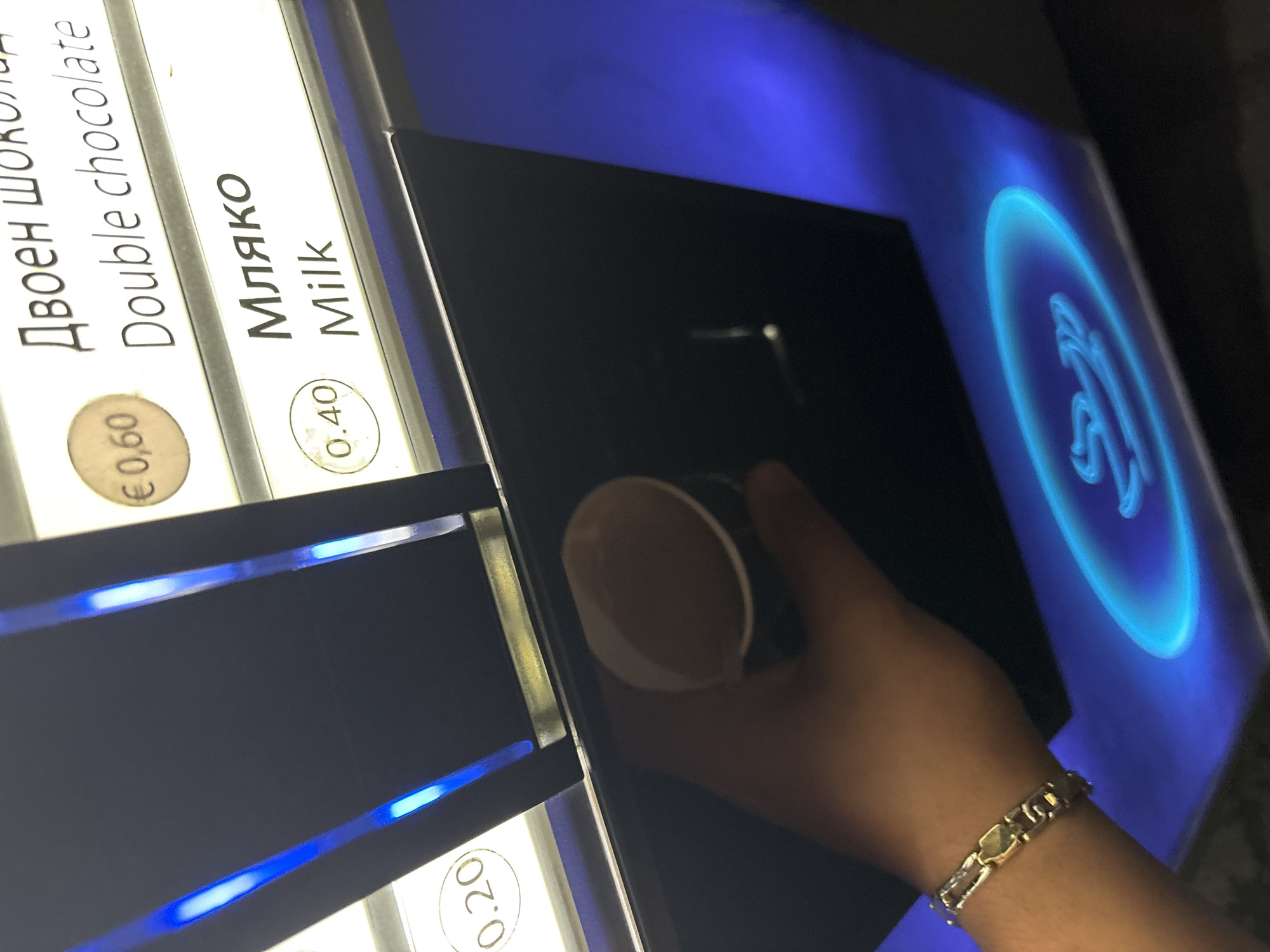 A person's left hand is holding a paper cup of coffee in front of a vending machine. The machine has a dark blue, black, and white interface with illuminated text and prices in Euros. Above the cup, the vending machine displays options such as "Double chocolate" for €0.60 and "Milk" for 0.40. The hand is adorned with a gold bracelet. The background of the vending machine is illuminated with blue light. This scene appears to be in a public space, possibly a rest stop or a commercial area, and the presence of Bulgarian text on the machine suggests it is located in Bulgaria, potentially in the city of Provadia.