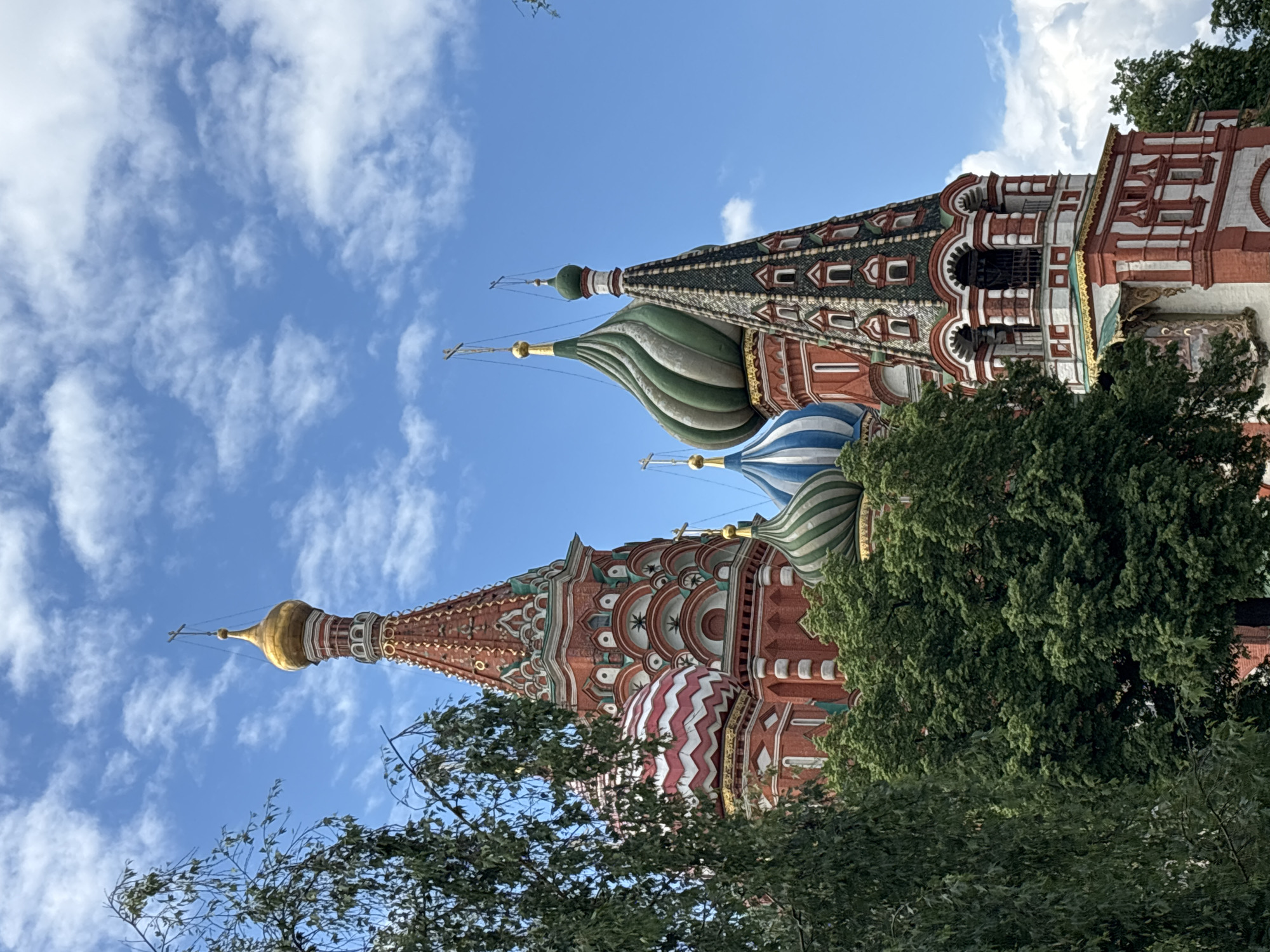 The image depicts an upward-facing view of St. Basil's Cathedral in Moscow, Russia. The iconic, colorful domes and spires of the cathedral are visible against a backdrop of a blue sky dotted with white clouds. Green trees partially obscure the lower portion of the building. The architectural details of the cathedral, including red brickwork, intricate patterns, and various colors, are prominent.