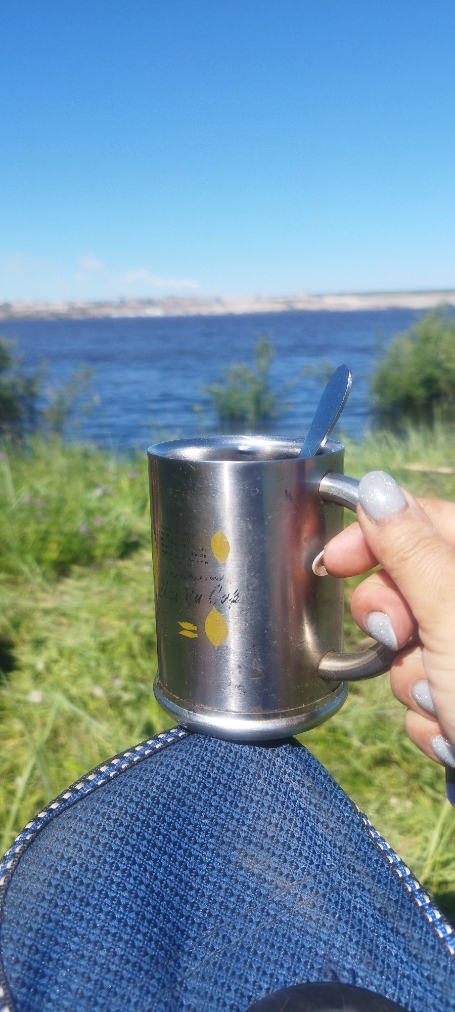 A first-person perspective shot shows a hand holding a metal mug with a spoon inside. The mug has two yellow lemon-shaped designs on it. The background is a body of water with a distant shoreline under a clear blue sky. The foreground shows a blue textured fabric, possibly clothing. The scene is outdoors, with green grass and some plants visible. The location is identified as Mokhsogollokh, Russia, suggesting a natural or recreational setting.