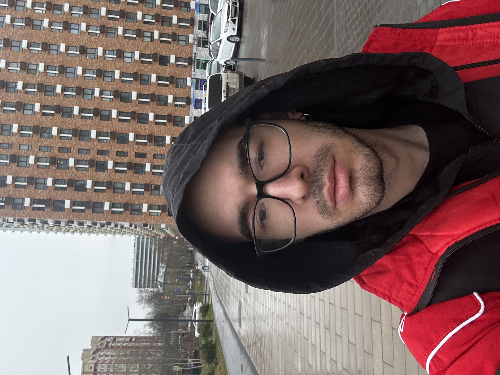 A person in a red jacket and black hooded sweatshirt is standing on a sidewalk in Moscow, Russia. The person is wearing glasses. In the background, there are buildings, including a tall residential building with a repeating pattern of windows. Cars are parked along the street, and the sky is overcast. The sidewalk is paved with rectangular blocks.