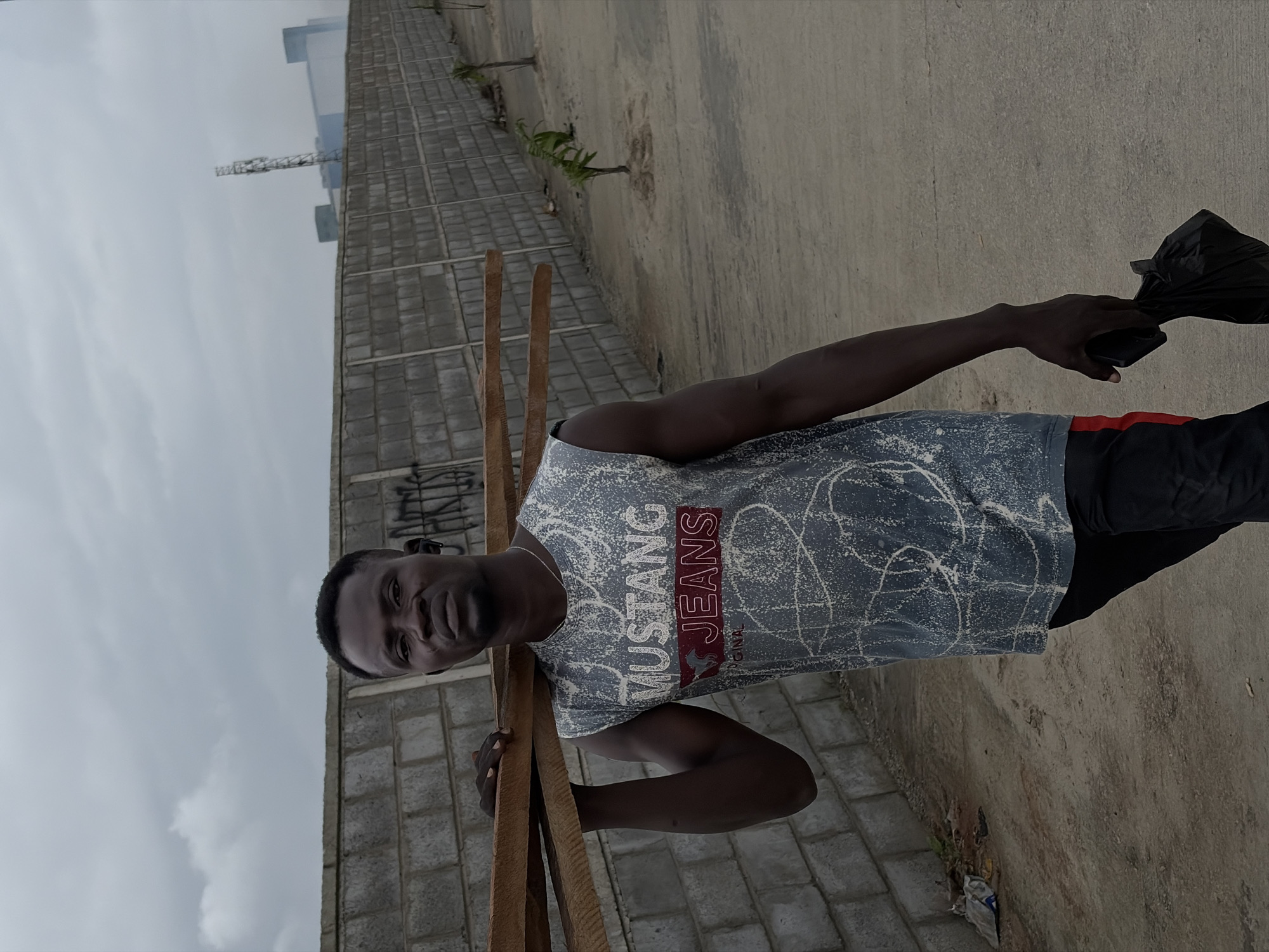 A dark-skinned man is pictured outdoors in Igboefon, Nigeria, carrying a stack of wooden planks on his left shoulder. He is wearing a grey, sleeveless t-shirt with a white, splattered pattern and the words "MUSTANG JEANS" in red and white text. He also has on black pants with a red stripe down the side. In his right hand, he holds a black plastic bag. The man is walking on a dirt and concrete path, with a grey cinder block wall to his left, which has graffiti on it. Beyond the wall, distant buildings and a cell tower are visible under a cloudy sky. Several small, spindly trees are planted along the path to the right of the wall. The ground is uneven and strewn with some debris.