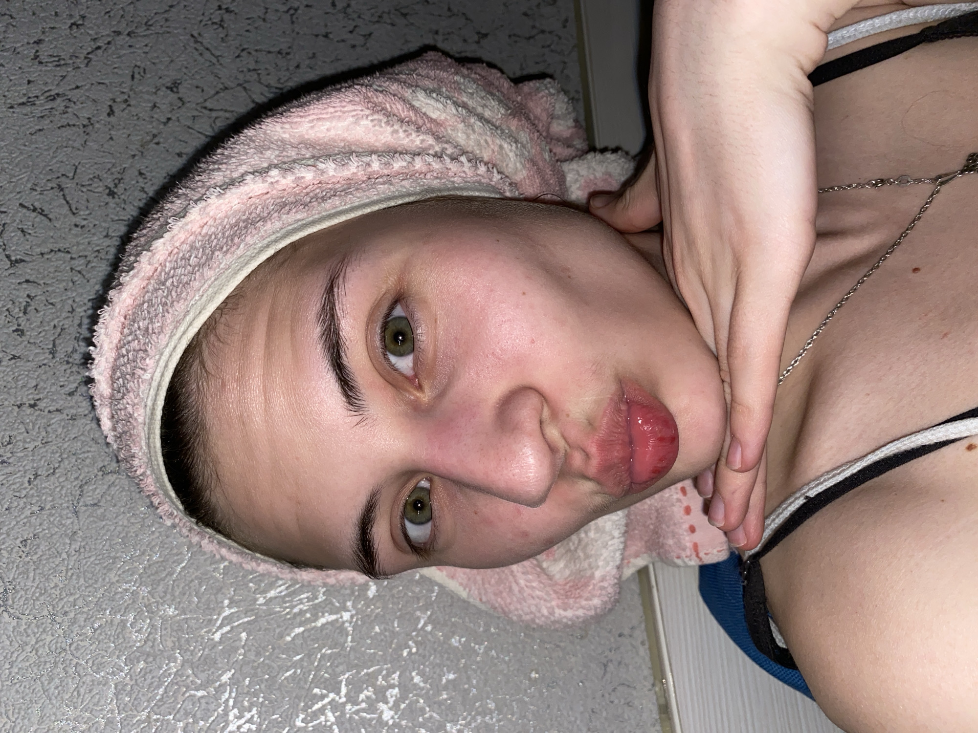 A young adult woman is lying on a textured gray surface, possibly a floor or bed, with a light pink and white striped towel wrapped around her head. She is looking at the camera with a slightly puckered expression. Her eyes are hazel. A silver chain necklace is visible against her skin. The woman's hand is near her neck, with her fingers slightly touching her throat. Part of a black and white garment (potentially a bra or tank top) is visible on her upper chest. The setting is simple, with no clear indication of being located in Votkinsk, Russia. No other information.