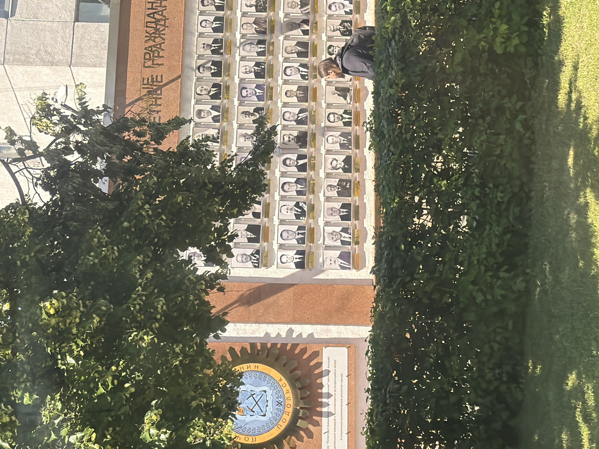The image shows a memorial wall in Podolsk, Russia, dedicated to "Honored Citizens." The wall features rows of framed portraits of individuals, with the inscription "ПОЧЕТНЫЕ ГРАЖДАНЕ" (Honored Citizens) prominently displayed at the top. To the left of the portraits is a circular emblem with the words "ПОДОЛЬСК" (Podolsk) around it. Below the emblem, a plaque is visible with Cyrillic text. A person wearing a dark jacket and backpack is walking in front of the memorial wall and a manicured hedge. The scene is outdoors, with sunlight casting shadows on the wall and the grassy area in the foreground.