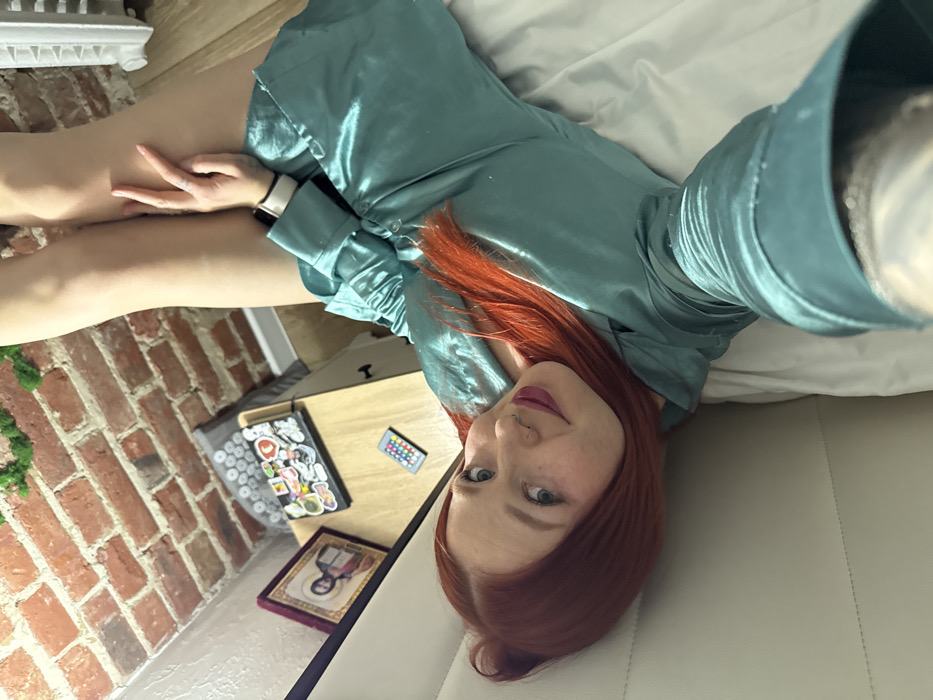 A female individual is positioned supine on a bed in a domestic interior setting, located in the city of Rostov-on-Don, Russia. She has long, reddish-orange hair and is wearing a teal-colored satin long-sleeved shirt. Her head is turned towards the camera, displaying a direct gaze, a slight smile, and visible freckles; she wears dark lipstick and a nose piercing. Her right hand rests near her hip, fingers splayed, and a white smartwatch is on her left wrist. Her bare legs are bent at the knees in the upper right quadrant of the frame. The bed features a light-colored, padded headboard and white bedding. The background wall is constructed of exposed reddish-brown brick, with green moss-like decorative elements affixed to it. A light-colored wooden nightstand with two dark drawer pulls is situated against the brick wall. On the nightstand, a framed Orthodox icon, a black laptop/tablet covered in stickers, and a small, multi-colored button remote control are visible. A grey acupressure mat is partially visible behind the laptop. A white, vertically-ribbed radiator is present on the far right. The floor consists of a light-colored wooden or laminate surface.