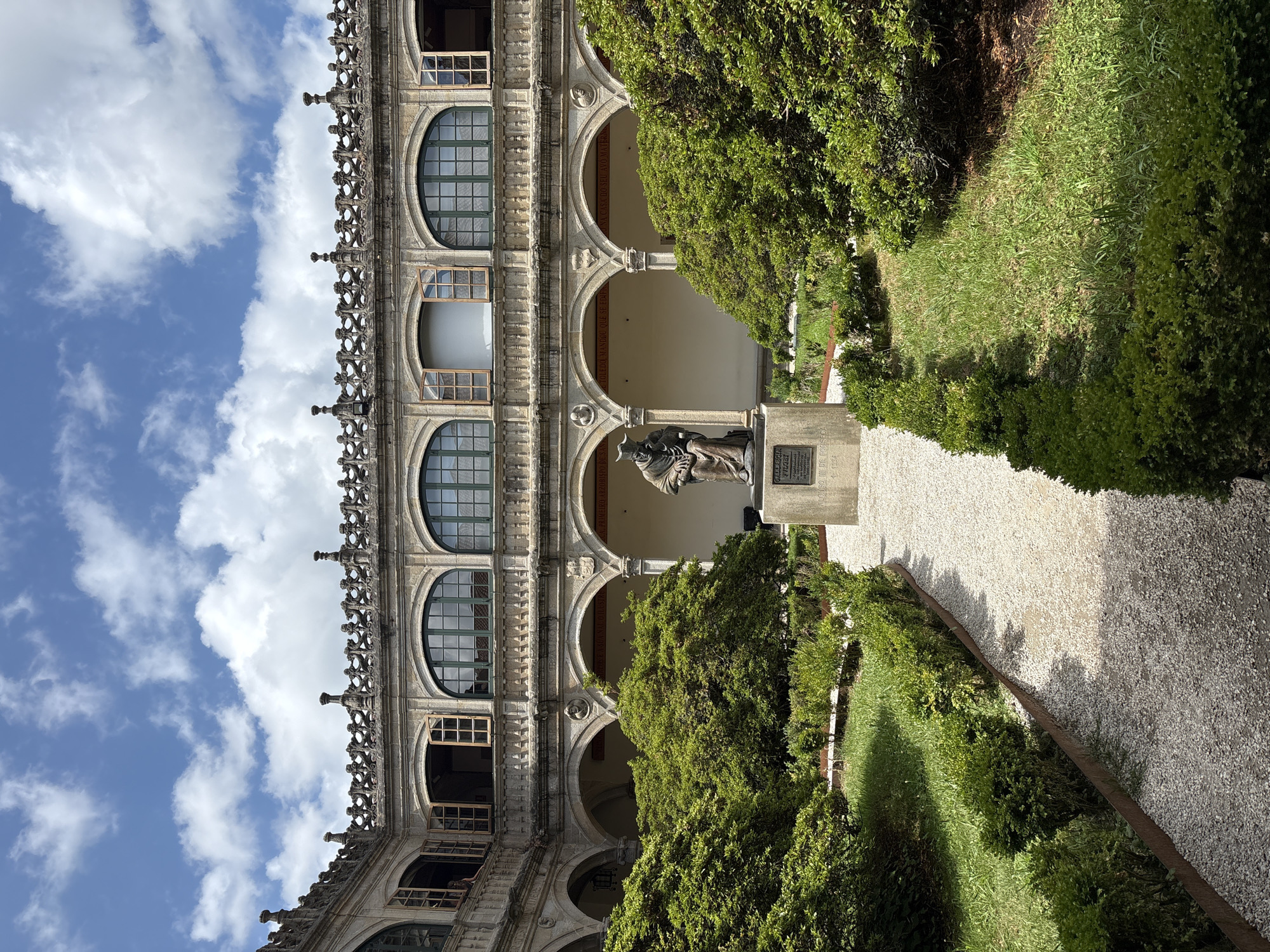 A courtyard in Santiago de Compostela, Spain features a statue of a cloaked figure seated on a pedestal in the center, surrounded by manicured green bushes and a gravel path. The courtyard is enclosed by a two-story building with arched walkways on the ground floor and arched windows on the upper floor. The building's facade is made of stone, and it has decorative elements along the roofline. The sky above is blue with scattered white clouds. The statue appears to be made of bronze and is located within a garden area, with the building forming the backdrop. The scene is well-lit by natural sunlight.