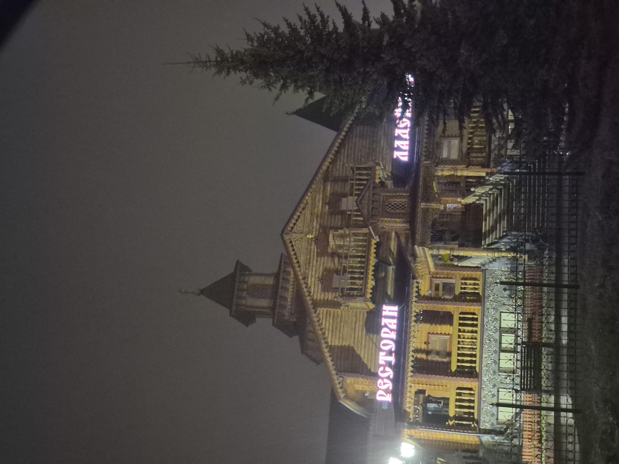 At night in Tobolsk, Russia, stands a large wooden building, likely a restaurant. A tall, multi-tiered structure with decorative carvings is illuminated, displaying signs in Russian that say "Ресторан" (Restaurant) and another word with the letters "AAAA" visible. Adjacent to the building is a dark evergreen tree. A metal fence runs along the property line. The sky is overcast and dark.