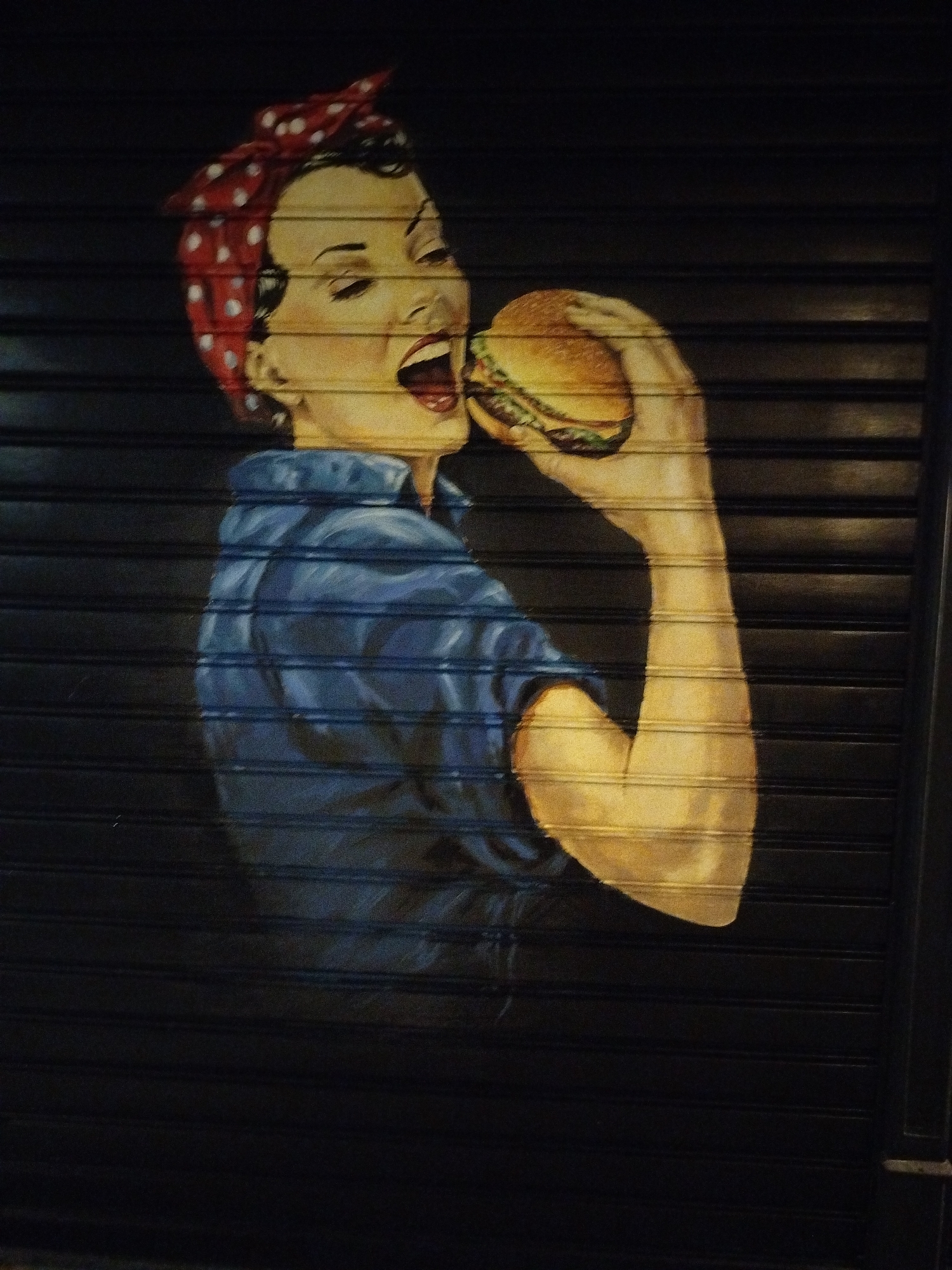 A painting on a dark, corrugated metal shutter in Novi Sad, Serbia, depicts a stylized woman in a red polka-dot bandana. She is wearing a blue denim shirt, and her right arm is raised, holding a hamburger to her open mouth, suggesting she is taking a bite. Her eyes are closed, and her facial expression conveys enjoyment. The painting is executed in a realistic style with visible brushstrokes, and the overall lighting is dramatic, highlighting the woman's face and arm against the dark background. The shutter's horizontal ridges are clearly visible through the painted surface.