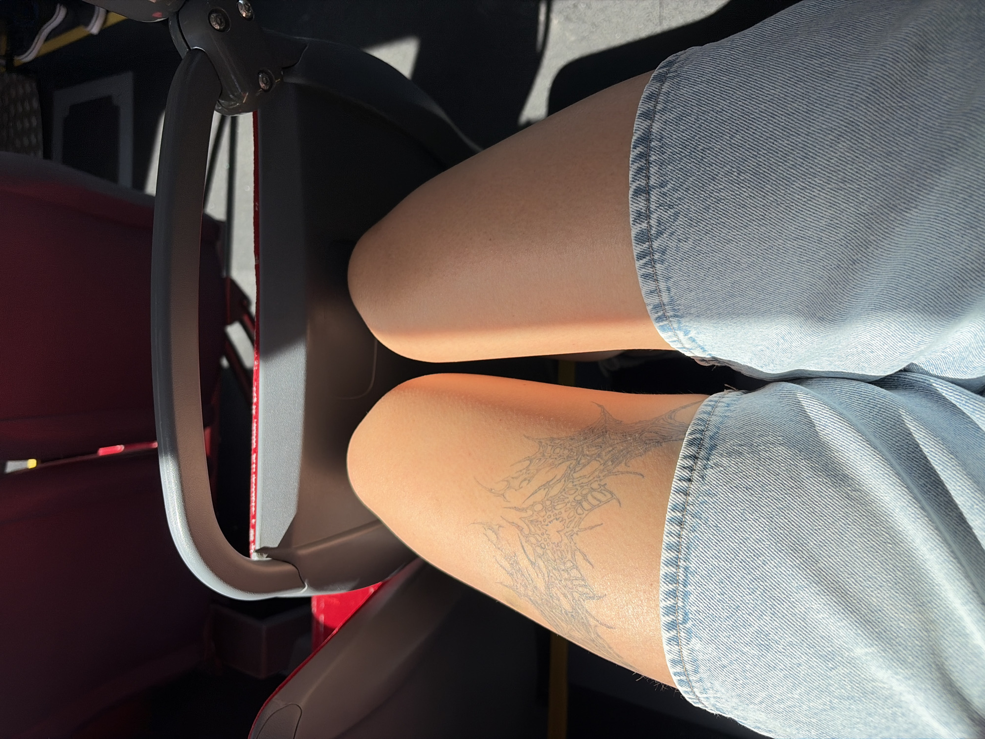The image shows a close-up view of a person's legs and thighs, wearing light-wash denim shorts. A tattoo, appearing to be a stylized design of thorny branches or a spiderweb, is visible on the left thigh. The person is seated on a red bus seat with a gray armrest in the foreground. Sunlight casts shadows on the legs and the interior of the bus. The scene is likely within the city of Nitra, Slovakia, given the context provided.