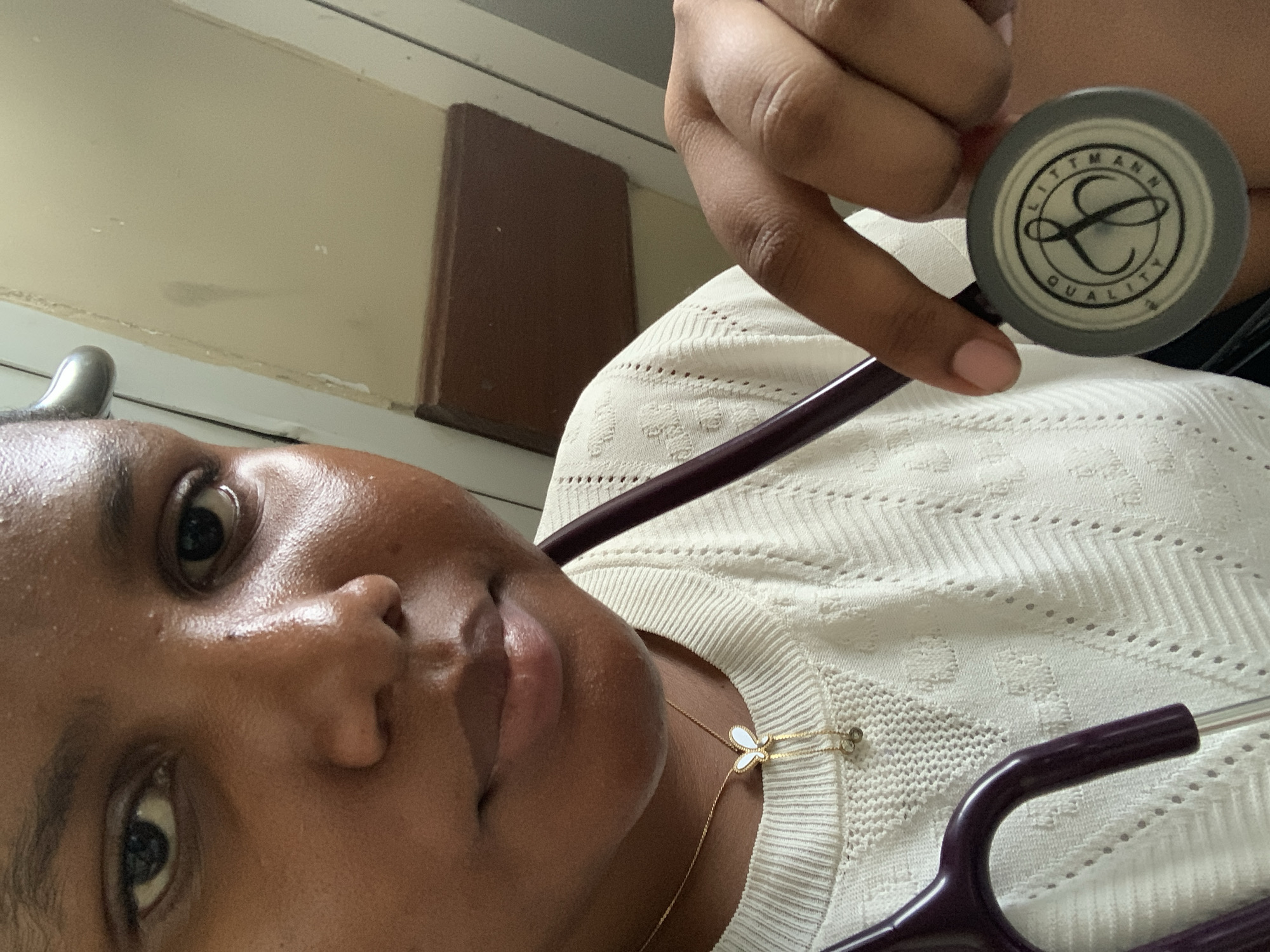 A person with dark skin holds a Littmann stethoscope near their chest while taking a selfie. The stethoscope is purple and gray. The person is wearing a cream-colored, patterned top and a gold necklace with a white butterfly pendant. The background includes a wall with a dark brown trim. The location is not discernible from the image.