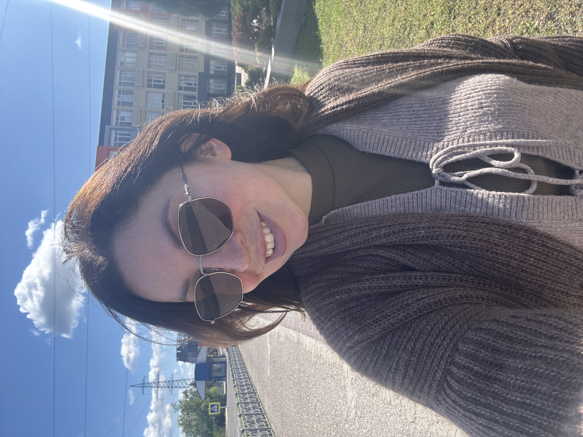 A young woman is captured in a vertical selfie, smiling broadly at the camera on a bright, sunny day in Kryvyi Rih, Ukraine. She wears dark, silver-framed aviator sunglasses and has long, dark brown hair. Her outfit consists of a dark olive green ribbed top beneath a light brown or taupe knitted cardigan, which features a decorative tie-front detail.
The setting is outdoors, likely on a bridge or overpass, with a patterned metal railing visible in the background. The sky is a clear blue with scattered white clouds, and a strong sun flare emanates from the upper left corner of the frame. In the distance, urban elements include several modern multi-story buildings and a large power pylon with overhead lines. Green grass and trees are also visible, suggesting a mix of urban and natural environments. A yellow diamond-shaped pedestrian crossing sign with a black figure is discernible in the lower left corner. The woman's cheerful expression and the bright sunlight convey a pleasant and happy mood.
