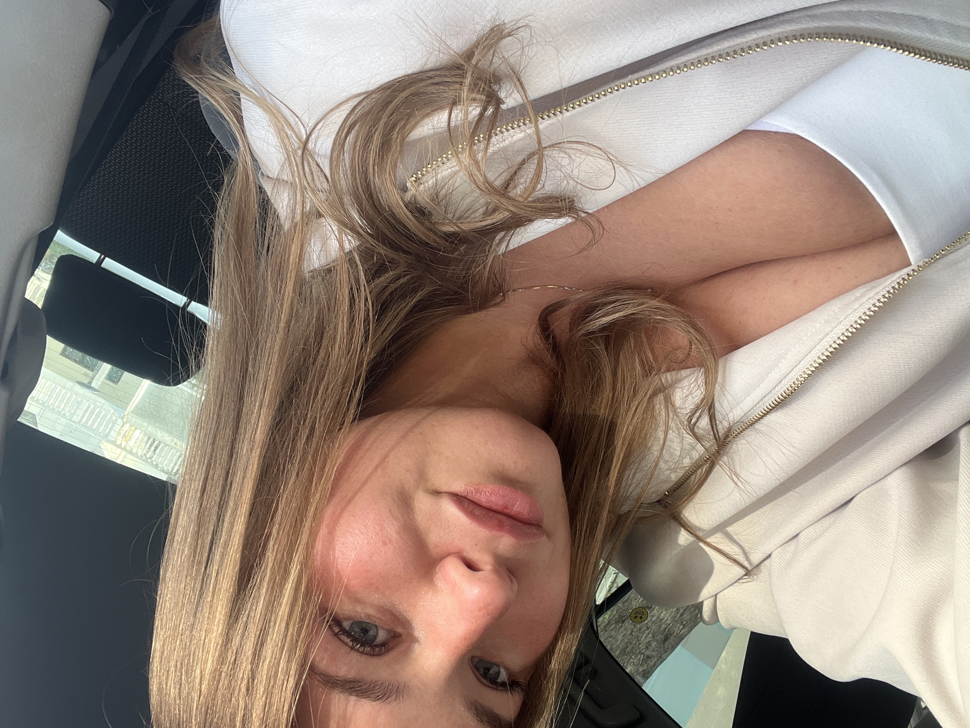 A woman with fair skin and light brown hair is in a car, likely the driver's seat, given the angle. She is wearing a white jacket with a gold zipper and a white shirt underneath. Her arms are crossed. The vehicle appears to be parked or stationary, based on the view through the car window, which shows a building across the street. Given the location of Budva, Montenegro, this could be a street scene within the city, although specific landmarks aren't visible to provide a pinpoint location. The interior of the car is dark, contrasting with the bright clothing and sunlight.