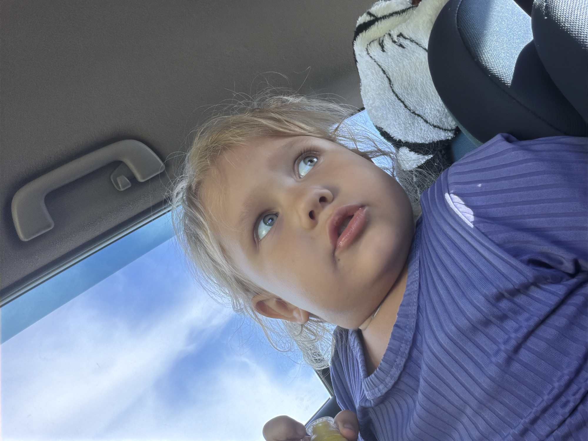 The media file shows a close-up, low-angle shot of a young child with blonde hair, looking upwards. The child is wearing a purple, ribbed shirt. The background is out of focus, but the sky with some clouds is visible through a car window, suggesting the child is in a vehicle. A grey car handle is visible on the left side of the frame, and a portion of a car seat with a patterned cover is on the right. The lighting suggests it is daytime, possibly late morning or early afternoon, with the sun casting shadows. The child appears to be holding a small, yellow object, possibly a bottle or food item, in their right hand. The overall scene is casual and appears to be a candid moment, possibly during a car ride. There is no visible text in the image.