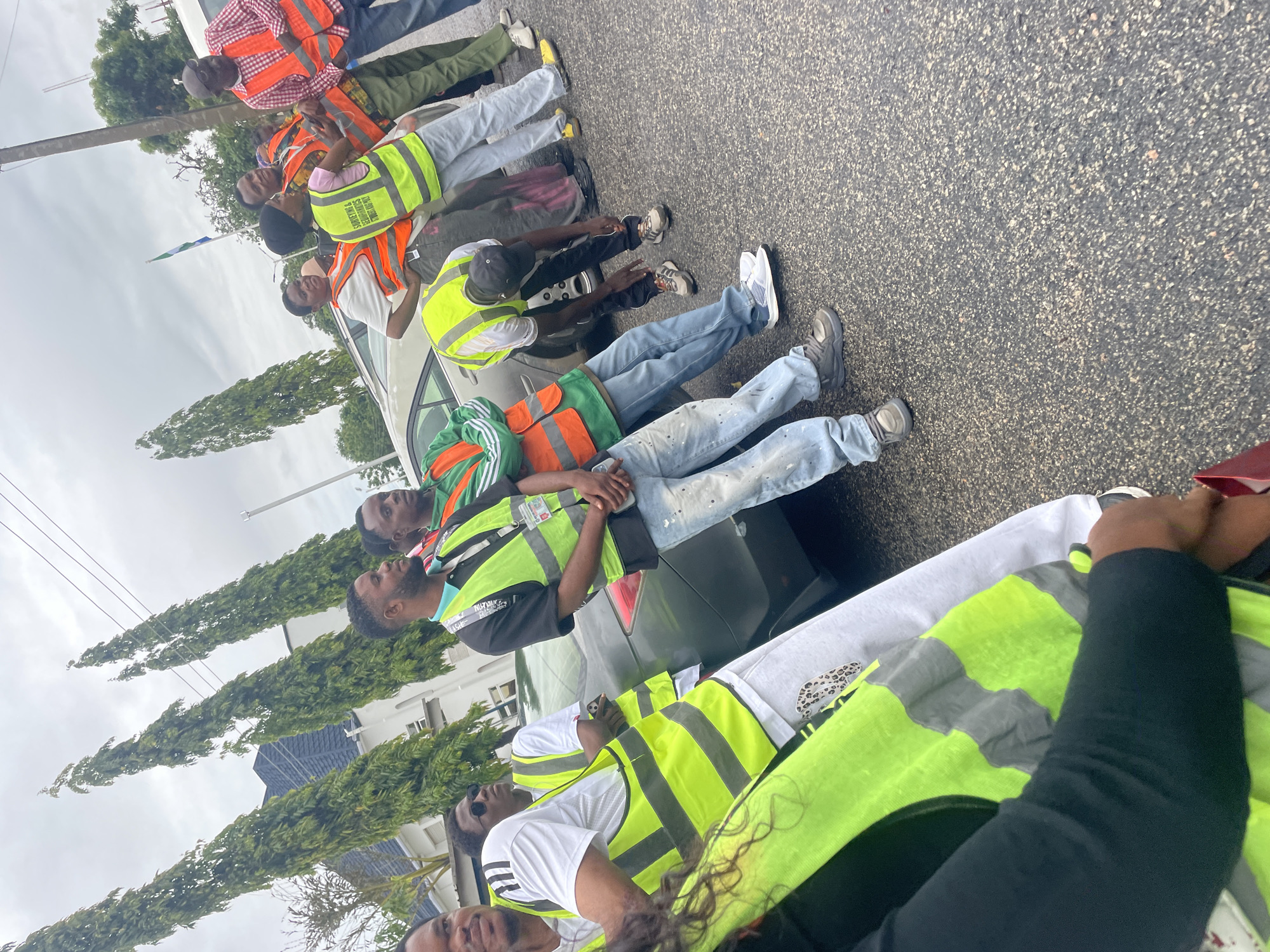 A group of people, appearing to be surveyors, are gathered on a paved street in Lokoja, Nigeria. Most are wearing high-visibility vests over their clothing. One person in the foreground is wearing a bright green vest with reflective stripes and has their arm in a sling. Several other individuals are wearing orange vests, and some are wearing green vests as well. They are standing near a dark-colored sedan. The environment includes tall, slender trees lining the street and a cloudy sky overhead. The individuals seem to be engaged in a work-related activity, with some looking towards the left and others interacting with each other.