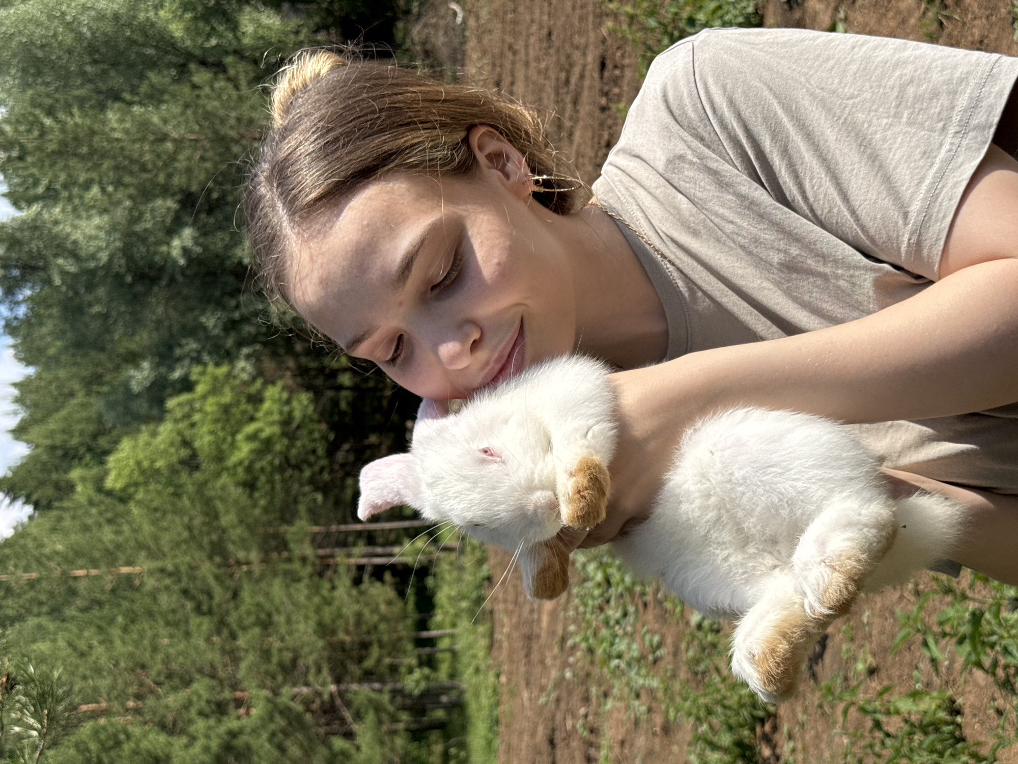 A young woman with light brown hair pulled back in a bun is holding a white rabbit with pink ears and red eyes close to her face. She is wearing a light brown t-shirt and a gold chain necklace with hoop earrings. The woman is outdoors in a wooded area with a field of brown dirt and green grass in the background. The trees in the background are tall and green. The overall scene suggests a warm, sunny day. The location is identified as Golyanovo, Russia.