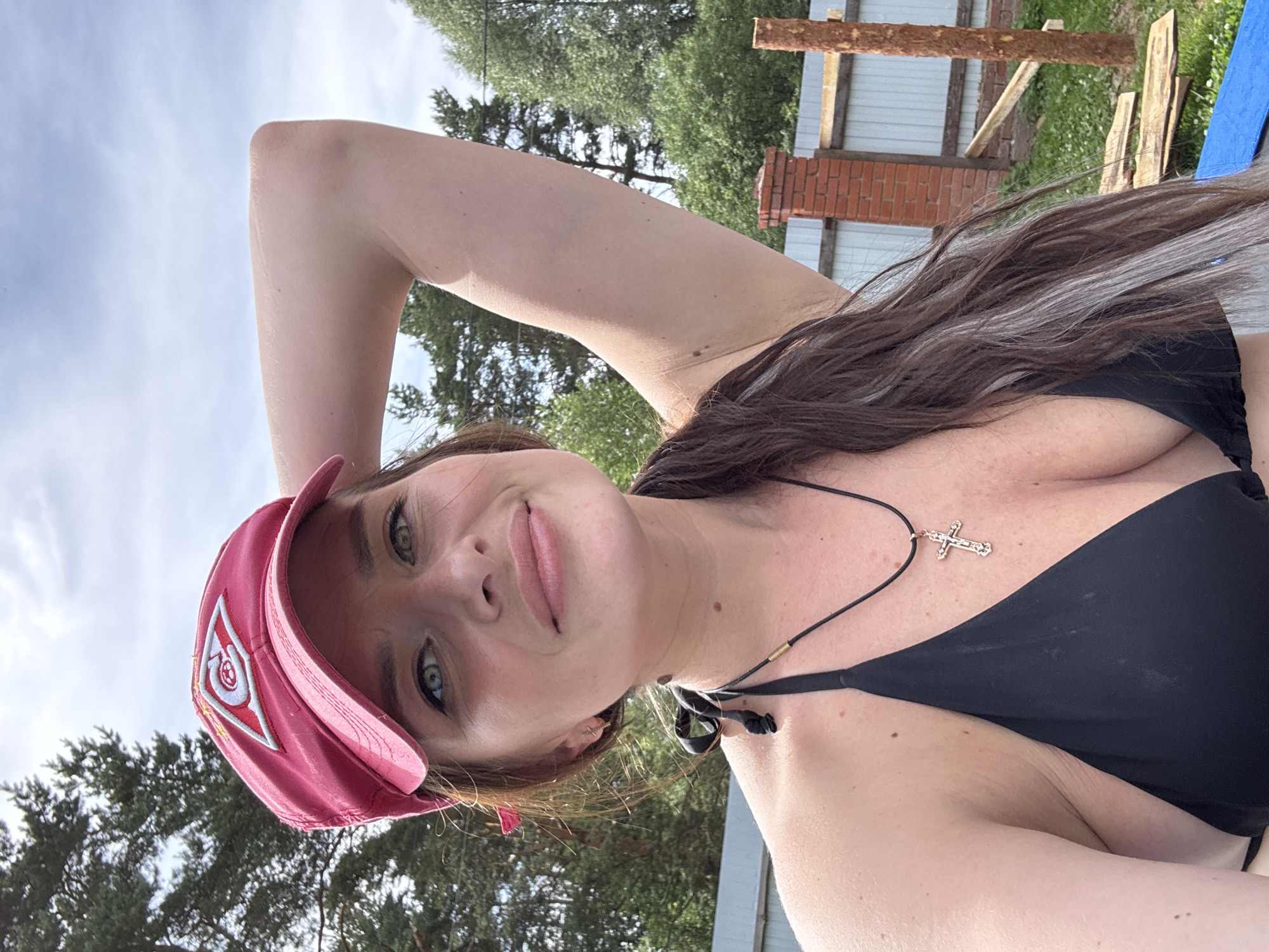 A woman is taking a selfie outdoors in what appears to be a rural or suburban area. She is wearing a red baseball cap, a black bikini top, and a necklace with a cross. Her right arm is raised, touching the back of her head. In the background, there is a wooden fence, trees, and some construction materials. The sky is cloudy. The image does not provide any specific information that would indicate the location in the city of Rusalevka, Russia.