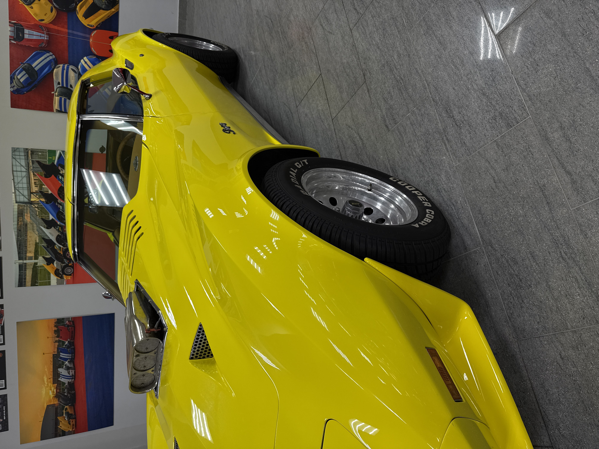 This media file, rotated 90 degrees counter-clockwise, captures a brightly painted, glossy yellow classic car, identified as a Chevrolet Corvette C3, likely a 427 model judging by the visible emblem on the fender. The car is showcased indoors, presumably within a showroom or museum in Sirius, Russia.
The primary subject is the vibrant yellow car, shown from a high-angle, rear-quarter perspective, highlighting its sleek lines and reflective surface. Notable details include chrome side mirrors, triple-outlet chrome side-exit exhaust pipes, and black mesh grilles on the bodywork. The car rides on large, multi-spoke silver wheels fitted with performance tires. Visible text on the tire sidewalls clearly reads "COOPER COBRA RADIAL G/T," with "GOODYEAR" partially visible on another tire.
The setting is an interior space with a clean, light grey tiled floor. The background features white walls adorned with posters depicting other classic cars and racing scenes, adding to an automotive theme. The lighting is bright and artificial, creating distinct reflections on the car's highly polished paintwork. No people or activities are visible, emphasizing the static display of the vehicle. The scene conveys a sense of meticulous presentation and preservation.