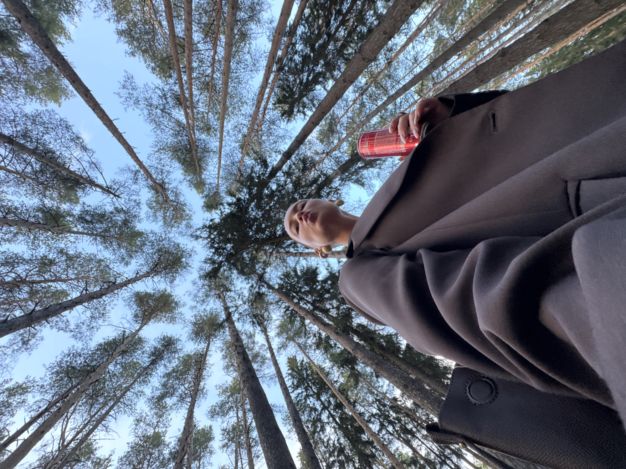 The image is a low-angle shot taken from below, looking up into a forest canopy of tall pine trees with a clear blue sky visible between the branches. The trees appear to be densely packed, creating a sense of enclosure. In the foreground, a person is visible wearing a dark-colored coat or jacket. The person is holding a red can, possibly a beverage, in their hand. The person's face is slightly blurred and out of focus, but their expression appears neutral. The overall impression is one of being in a forest setting, possibly a park or wooded area. Without more context, it is impossible to determine if the image was taken in Votkinsk, Russia.
