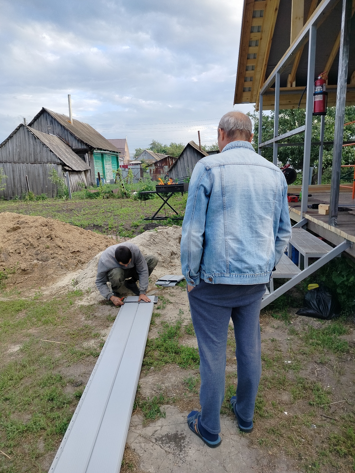 In Вазерки, Russia, two men are working outdoors near a partially constructed building. One man, wearing a blue denim jacket and grey sweatpants, stands facing away from the camera, observing the work. The other man, dressed in a grey sweatshirt and dark pants, kneels on the ground, measuring and marking a light grey siding panel with a ruler and what appears to be a utility knife. The siding is laid out on the ground. In the background, several weathered wooden structures, a pile of sand, and overgrown grass are visible. A metal frame for a building extension is also present, with a red fire extinguisher attached. The sky is overcast.