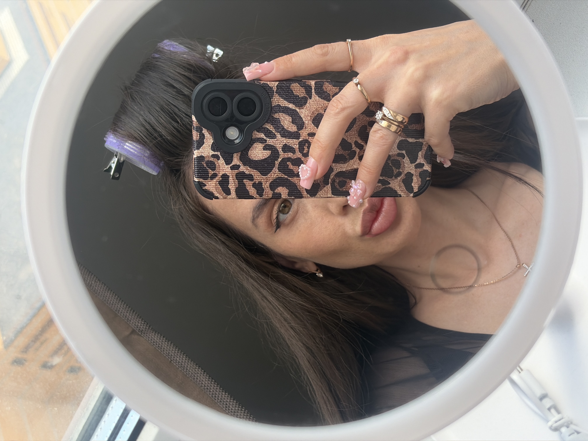 The image captures a mirror selfie of an adult female in an indoor setting within Novosaratovka, Russia. The subject, with long dark brown hair, has purple hair rollers secured by clips on the top and side of her head. Her right eye, featuring winged eyeliner, is visible, along with her full, slightly parted lips. Her right earlobe displays a small stud earring. She holds a smartphone with a leopard-print case and a prominent black multi-lens camera module, obscuring the left side of her face. Her right hand, adorned with several gold-colored rings, including one with a small clear stone, also displays long, pink fingernails with intricate 3D embellishments such as pearls and clear bows. A delicate gold-colored chain necklace with a small cross pendant is visible around her neck. The reflection is contained within a white-framed circular mirror, with a power cord visible at the bottom left. A blurred reflection of an outdoor scene, featuring brownish-orange structures, is visible in the top-left quadrant of the mirror, suggesting a window. A dark background, possibly a wall or interior upholstery, is visible behind the subject.