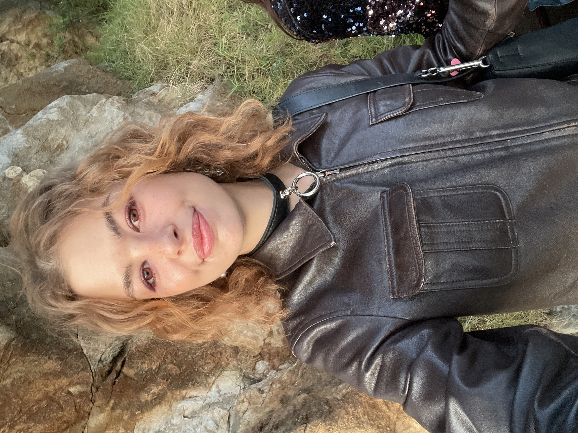 A young woman is captured in a close-up, rotated portrait, lying on her back in an outdoor setting. Her light, curly hair, possibly auburn or reddish-blonde, frames her face. She has fair skin adorned with prominent freckles across her nose and cheeks. Her light brown eyes are enhanced with red-toned eyeshadow, and she sports a subtle, closed-mouth smile.
She is dressed in a dark brown leather jacket featuring visible seams, a zipper, and flap pockets. Around her neck, she wears a black choker with a large, prominent silver O-ring and a small chain. A black shoulder bag with a silver clasp is visible, draped across her. In the upper right quadrant of the image, a glimpse of a black sequined garment or accessory can be seen.
The background consists of natural elements: rough, textured grey and brown rocks form the ground beneath and around her, interspersed with patches of dry, light green grass. The lighting is natural and soft, suggesting a clear day, possibly late afternoon or early morning. The overall mood is relaxed and calm, with the woman gazing directly at the viewer. There is no visible text. The rocky, natural landscape is consistent with an outdoor location in Budva, Montenegro.